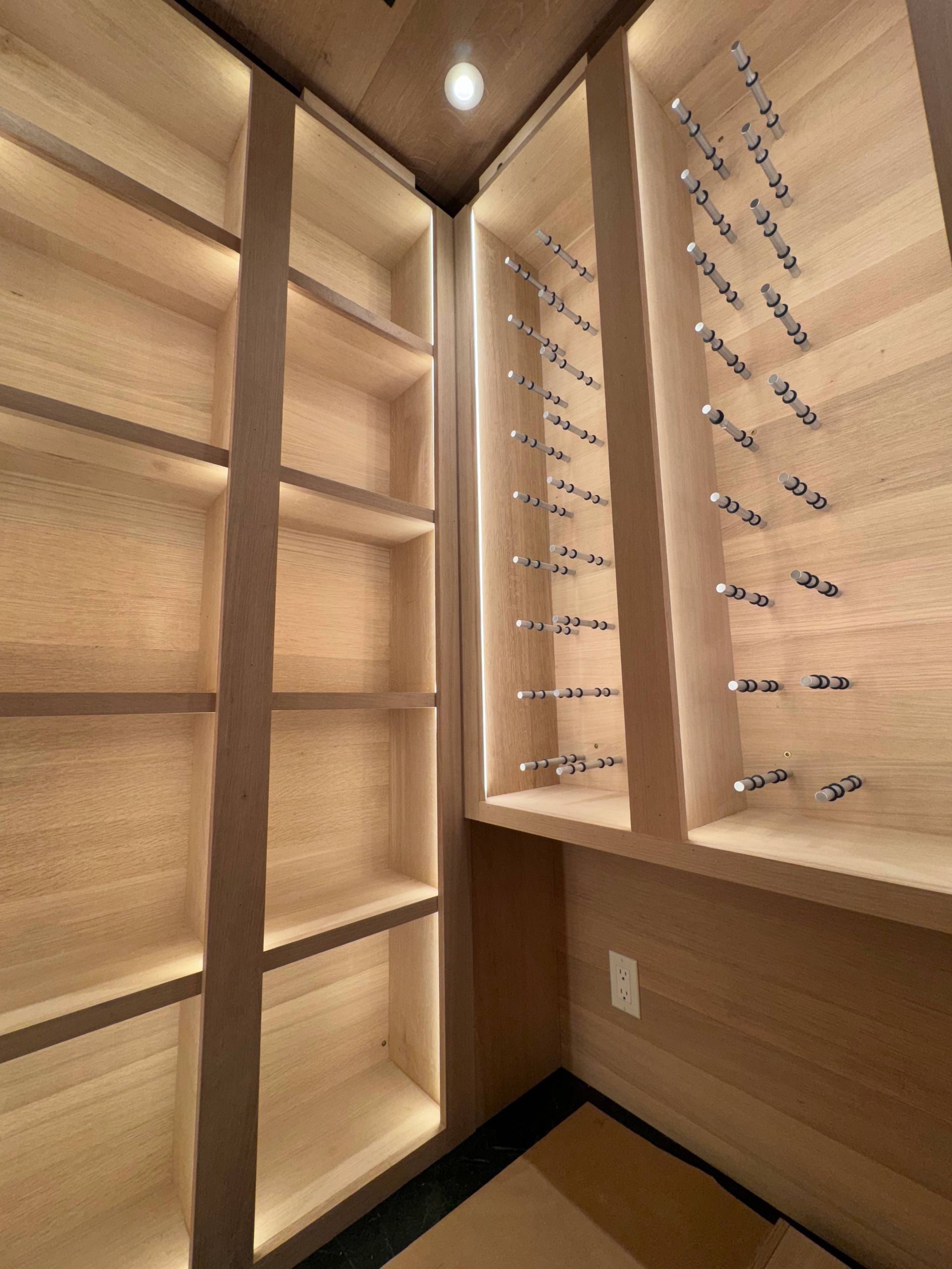 A corner wine cellar with light wood shelves, including a dedicated rack with metal pegs for individual wine bottles.