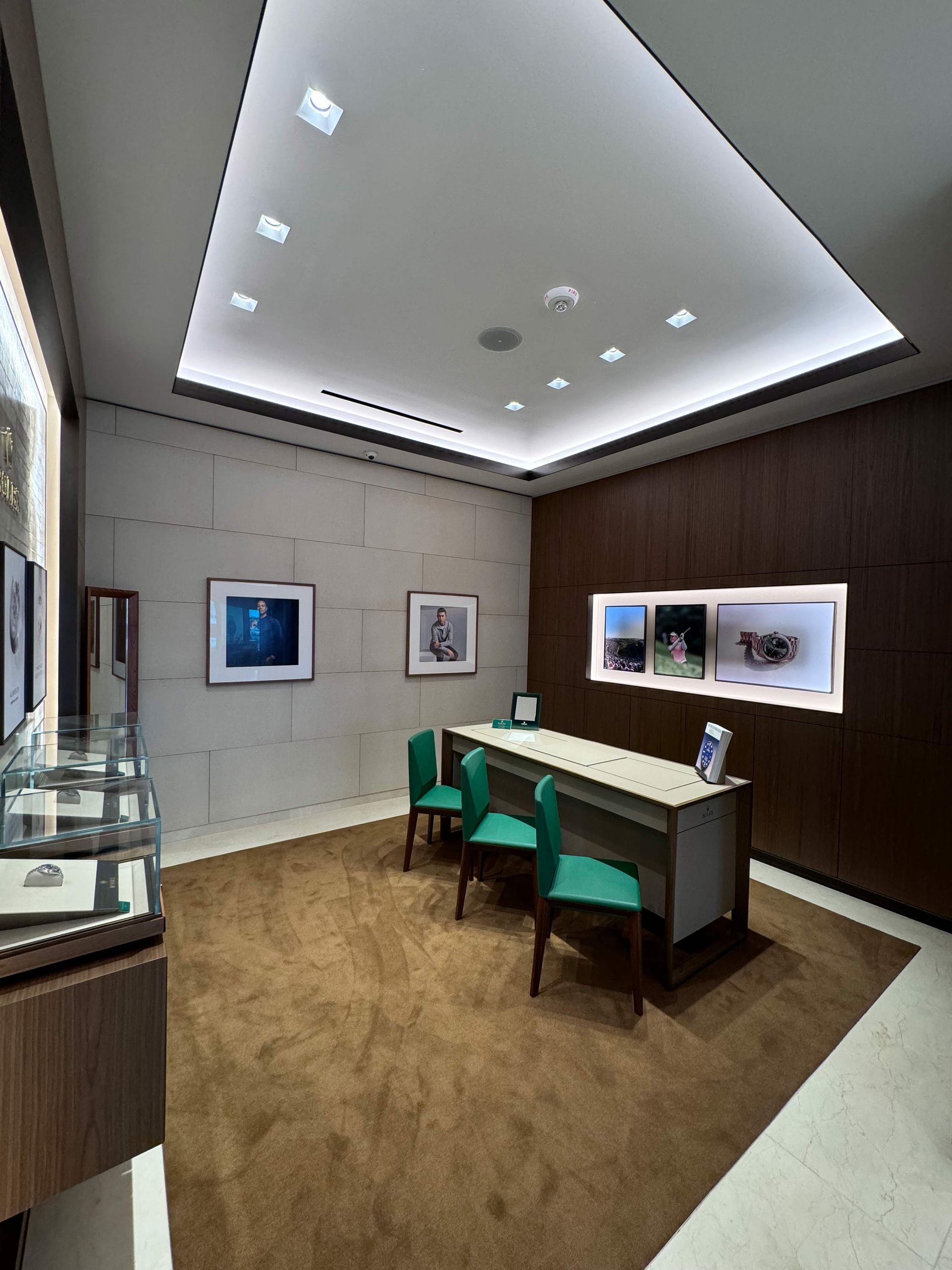 A modern office space with two green chairs at a desk, wood paneling, framed art, and recessed ceiling lighting.
