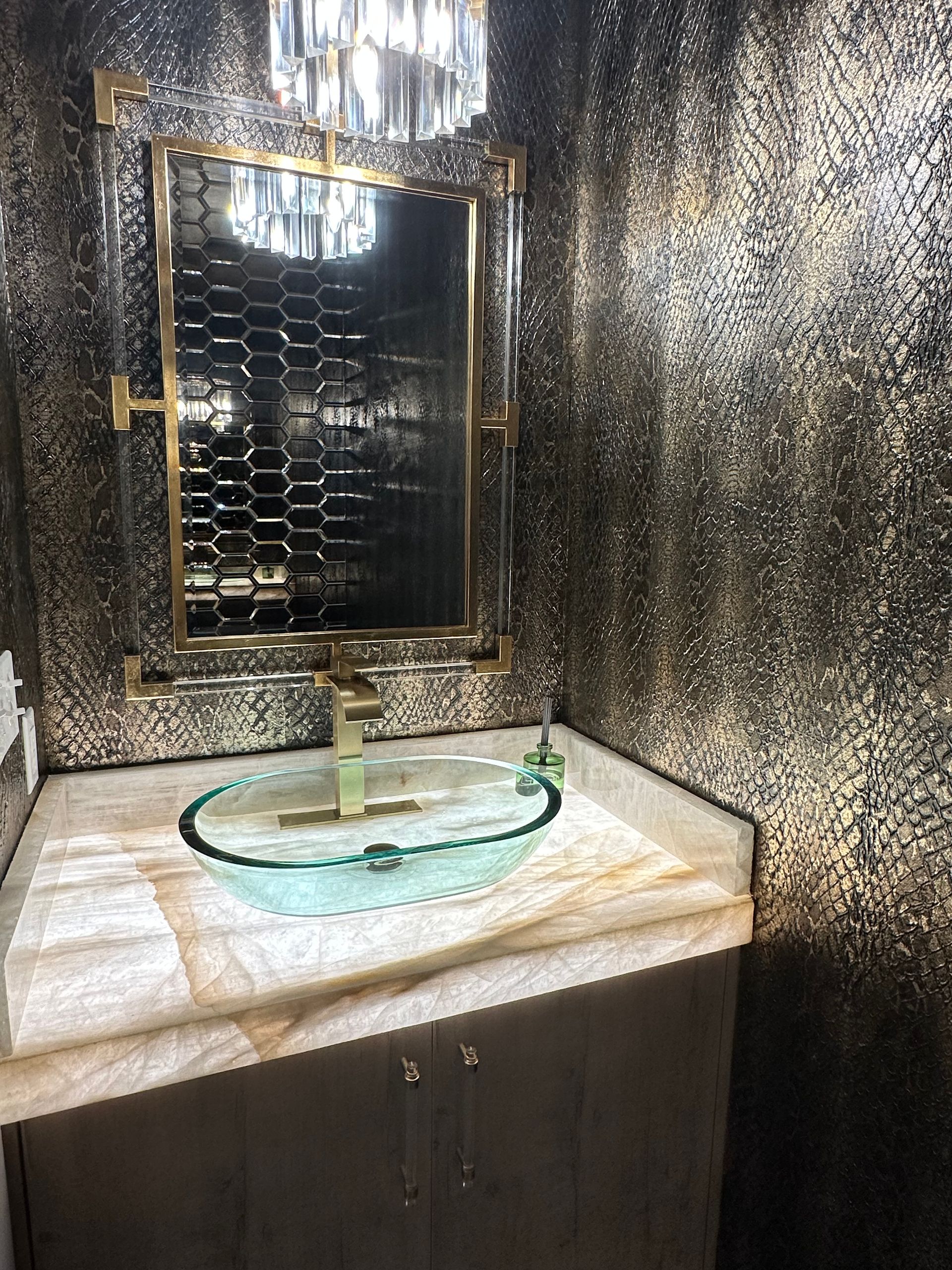 Bathroom with gold and black textured walls, gold fixtures, glass sink, and a decorative mirror.