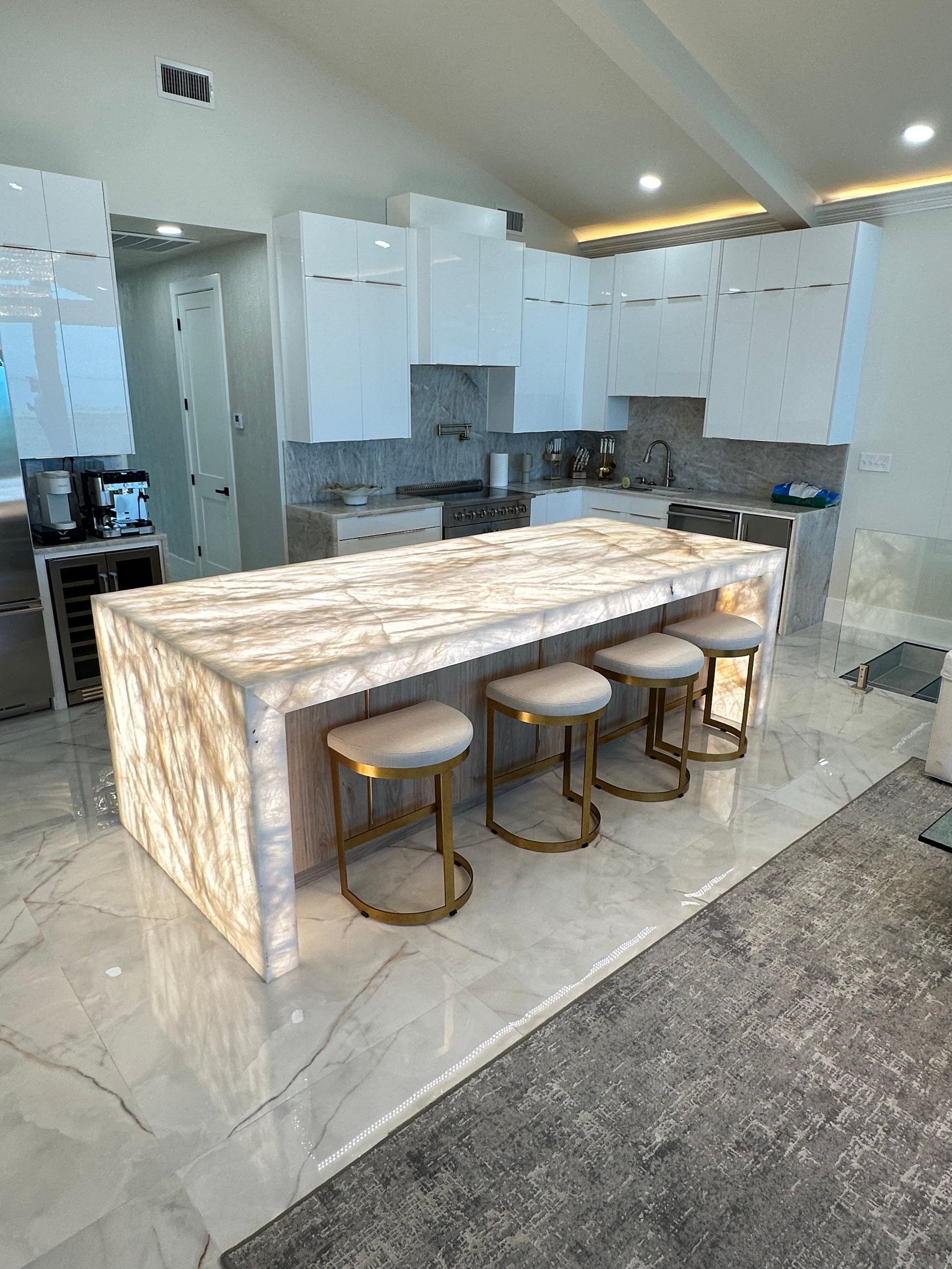 Modern kitchen with a lit marble island and four gold-legged bar stools. White cabinetry and marble counters are visible.