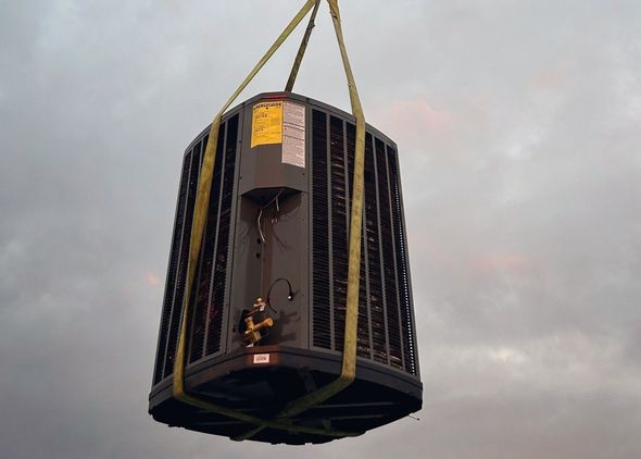 Air conditioning unit suspended by yellow straps against a cloudy sky.