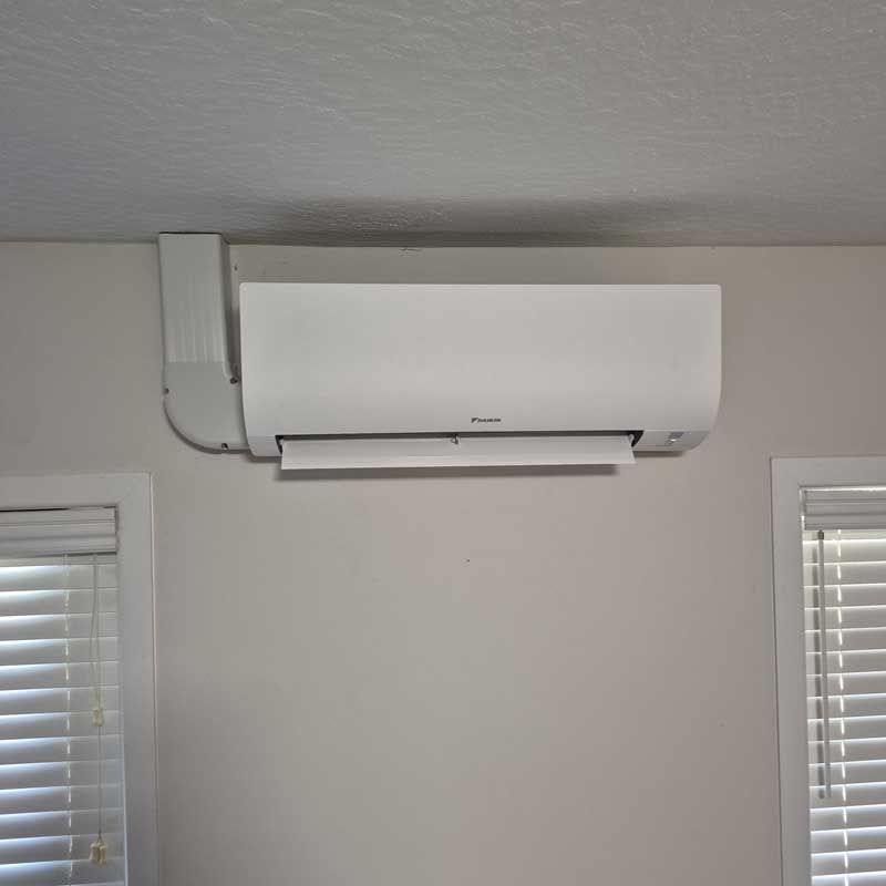 White ductless AC unit mounted on a wall between two windows with blinds.
