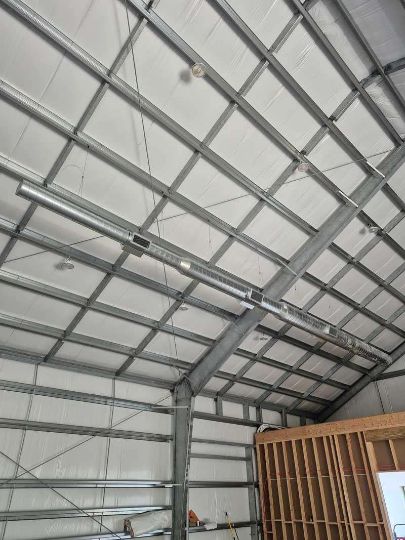 Interior view of a metal building's roof with insulation, beams, and a light fixture. A wooden framed wall is visible.