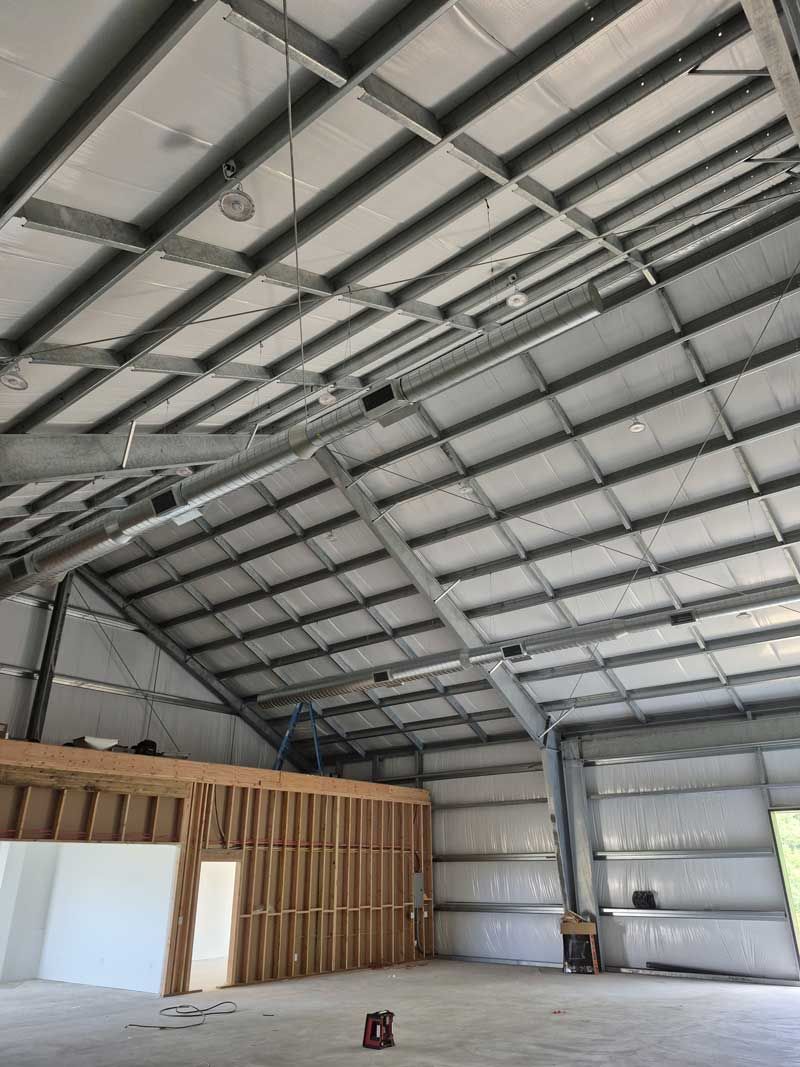 Interior of a large metal building under construction, with exposed steel beams and roofing. Wooden frame wall visible.