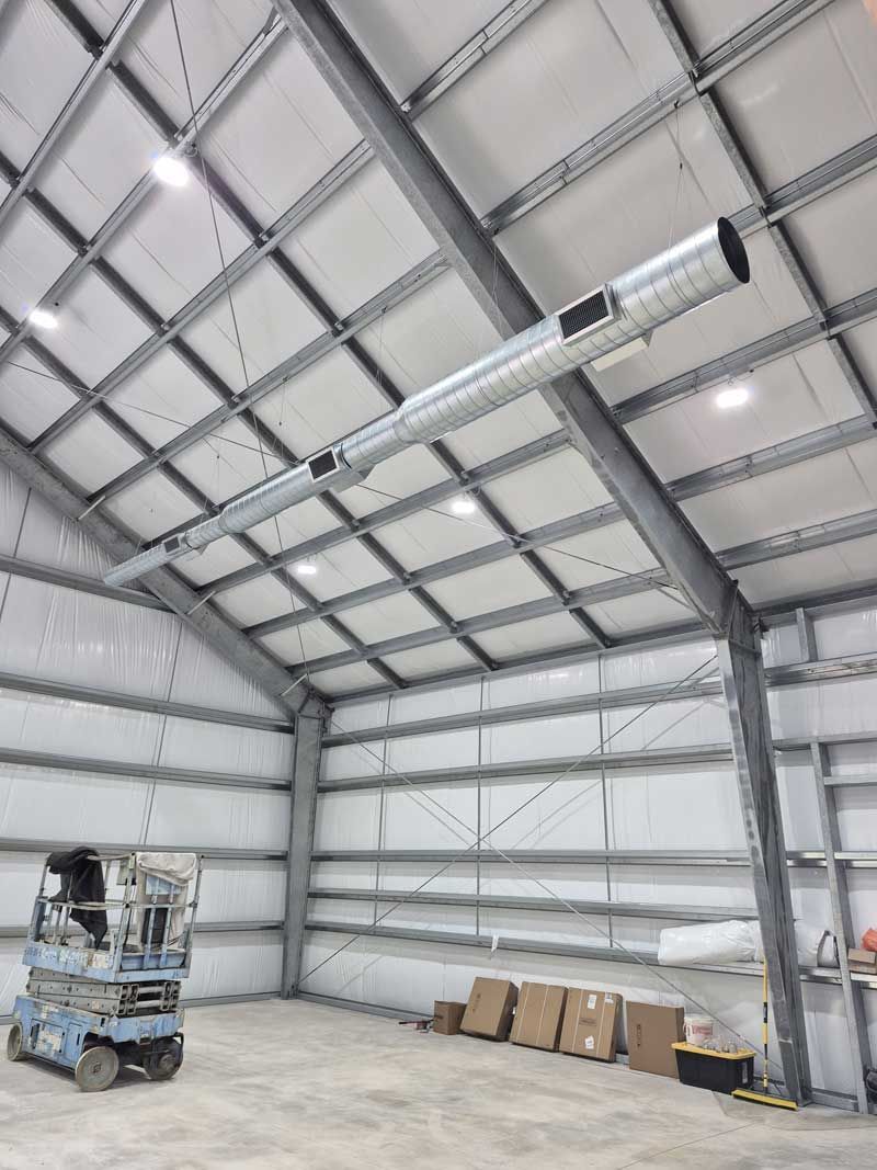 Interior of a metal industrial building with overhead lighting and ductwork. Boxes sit on the floor; a lift is on the left.