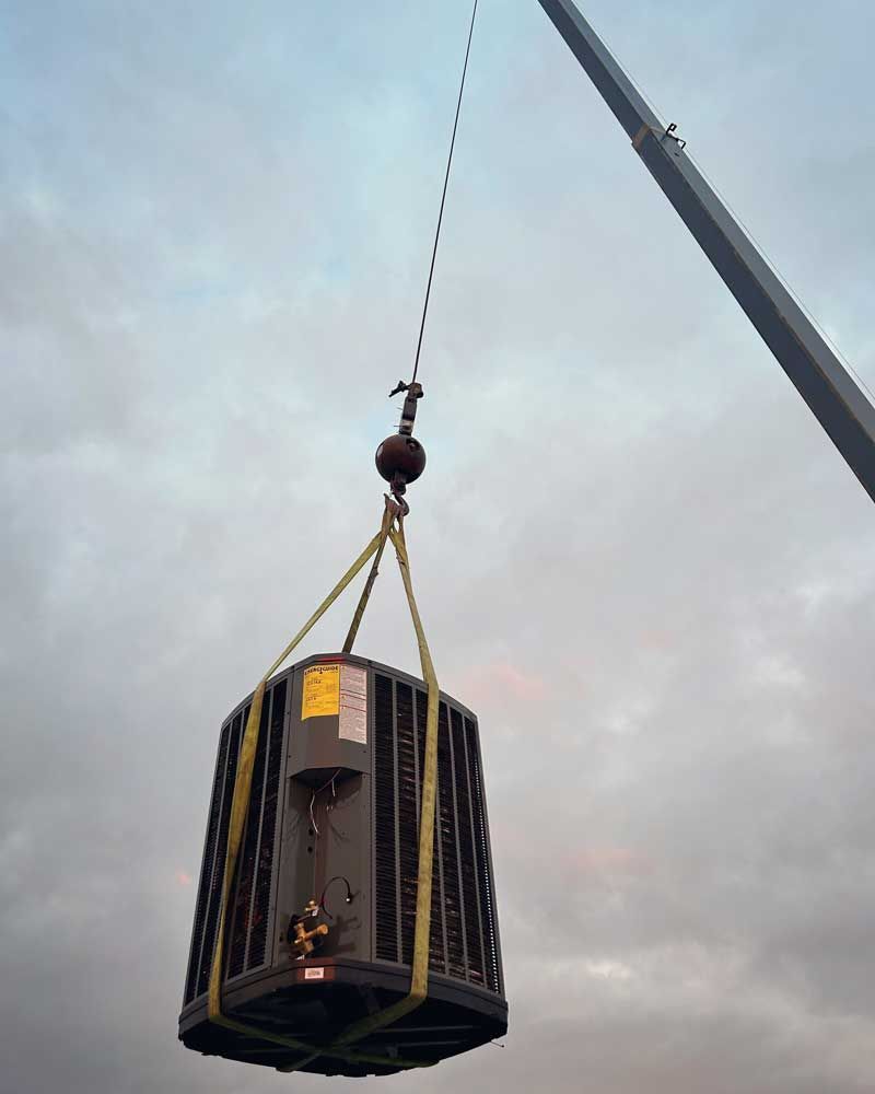 A large HVAC unit suspended by yellow straps from a crane against a cloudy sky.