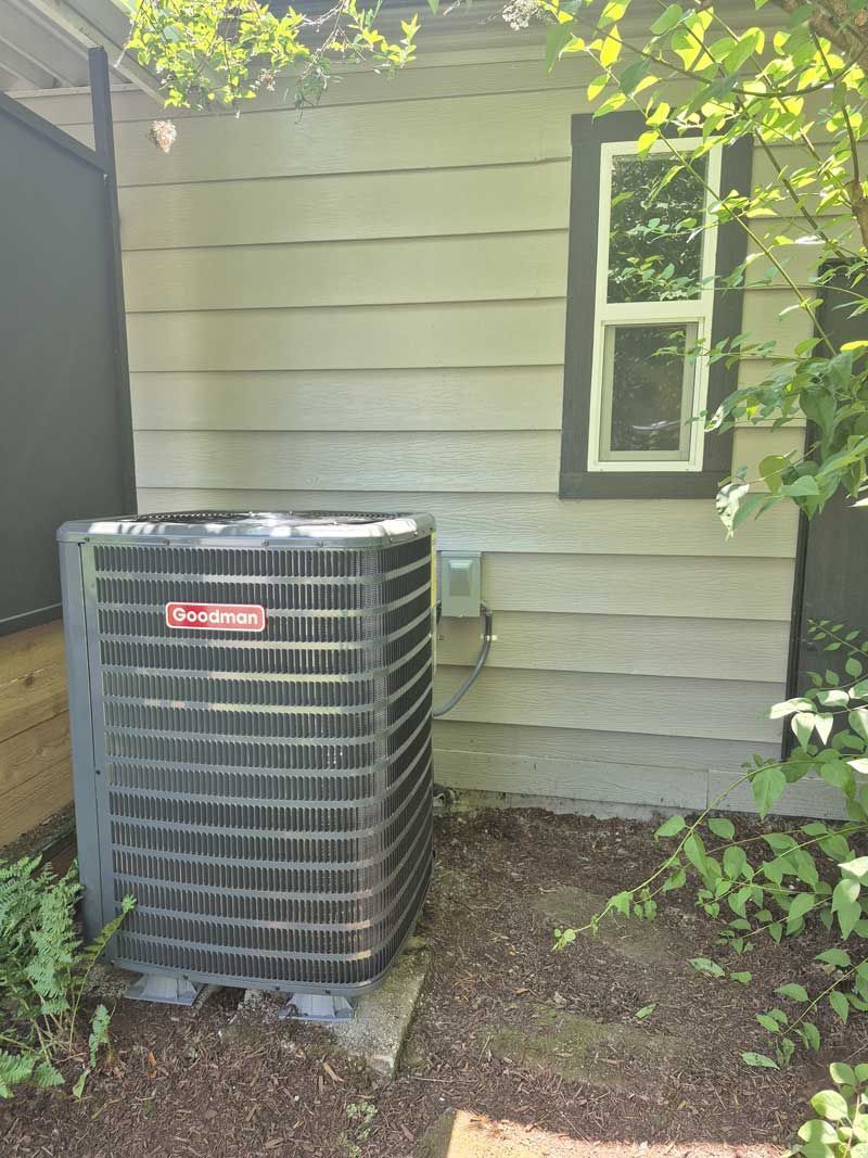 Gray Goodman air conditioning unit next to a light gray building with a window and vegetation.
