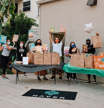 People holding up food and bags, smiling, behind a table with food and supplies, outside, HAVEN sign.