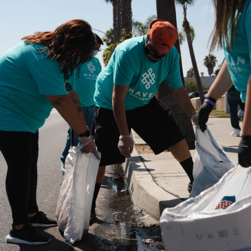 People in teal shirts picking up trash along a curb, placing it into white bags.