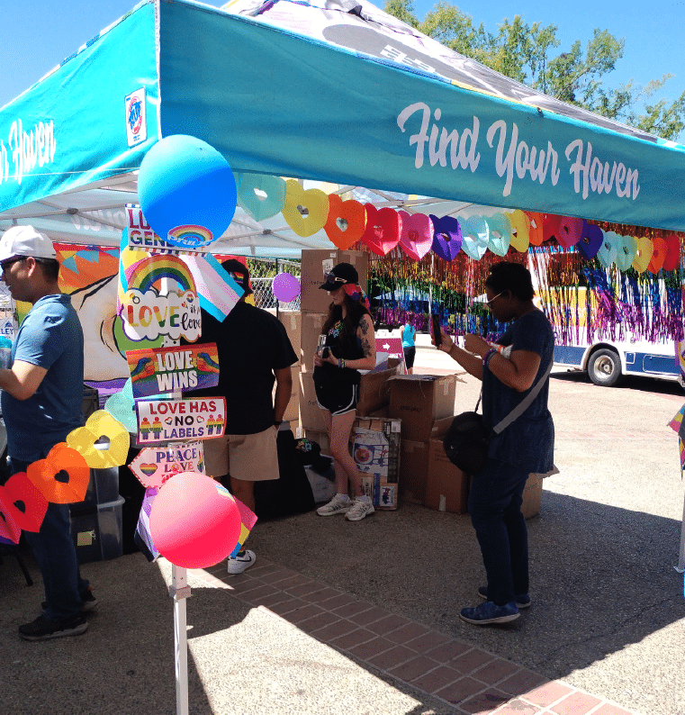 Pride event booth with rainbow decor. People gather, taking photos. 