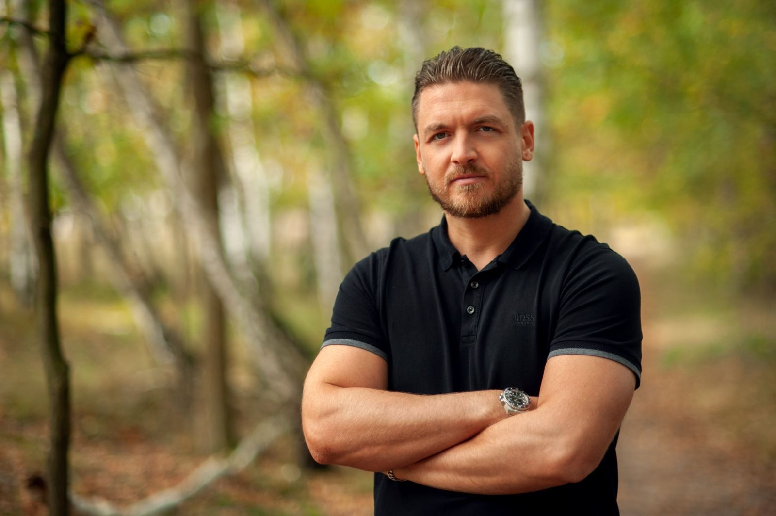 Man with crossed arms in black shirt, standing in a forest with blurred background.