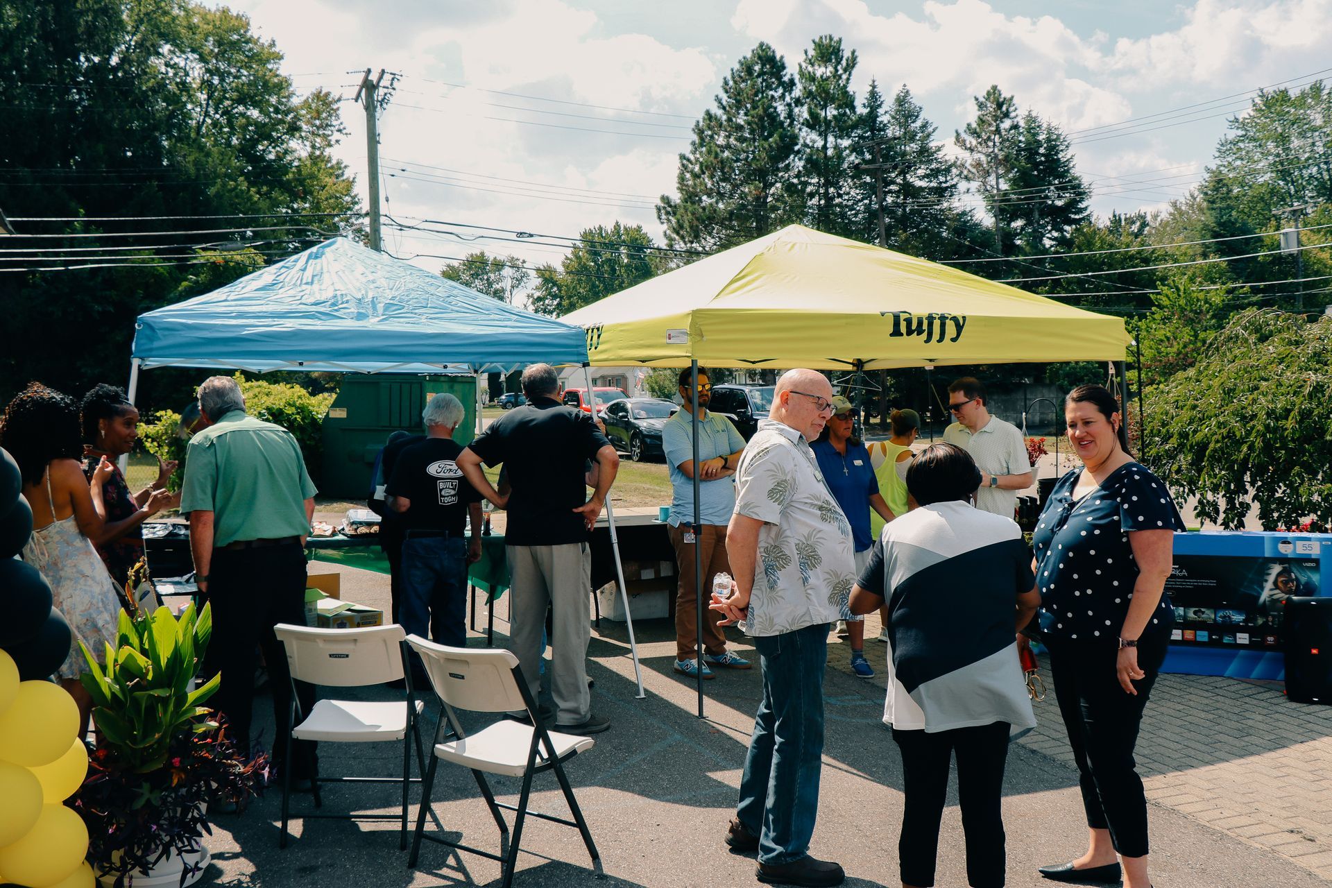 People gather outdoors near tents, some standing, some talking. Sunny day with trees and blue sky visible.