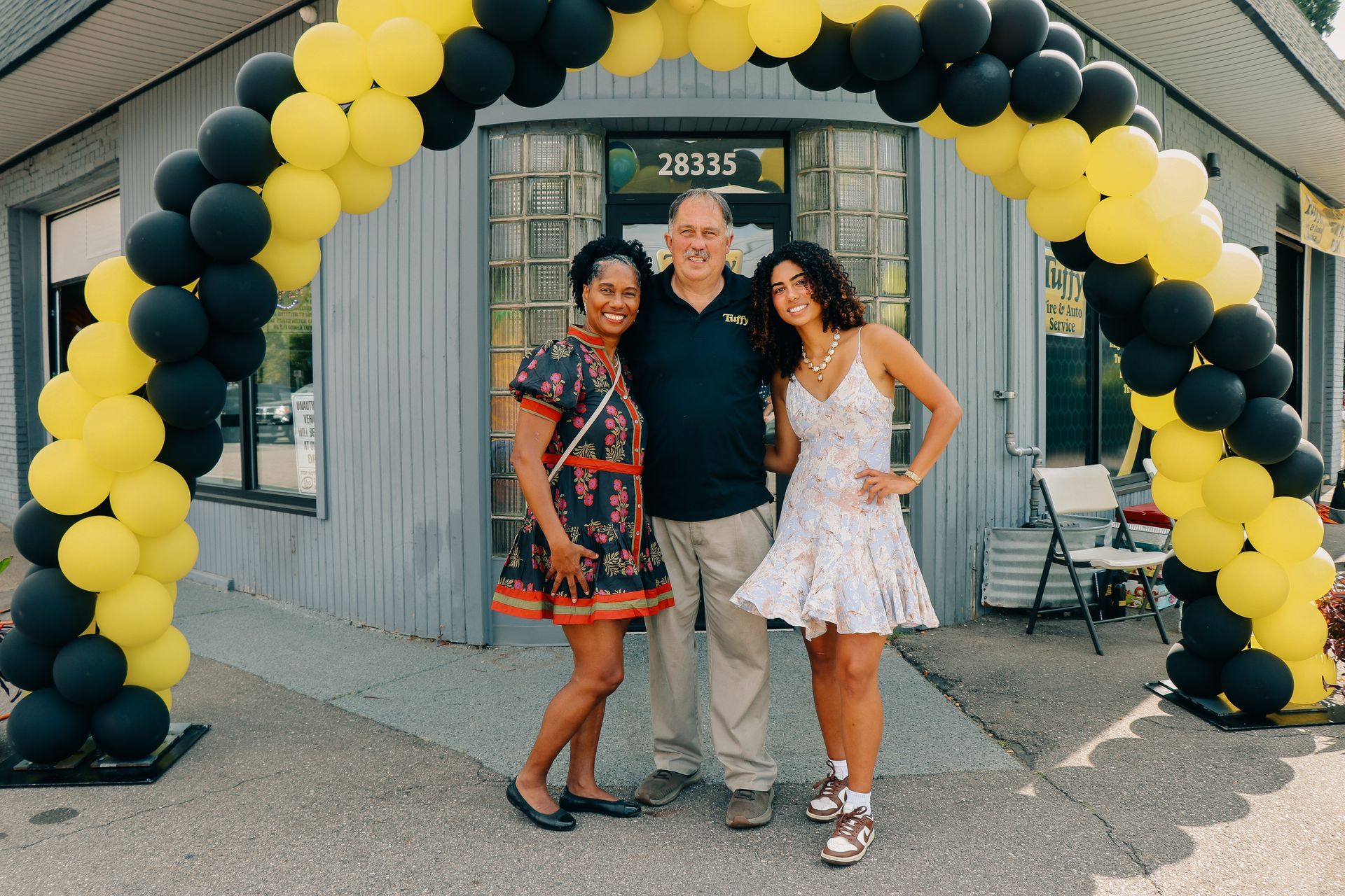 Three people pose under a yellow and black balloon arch in front of a building.