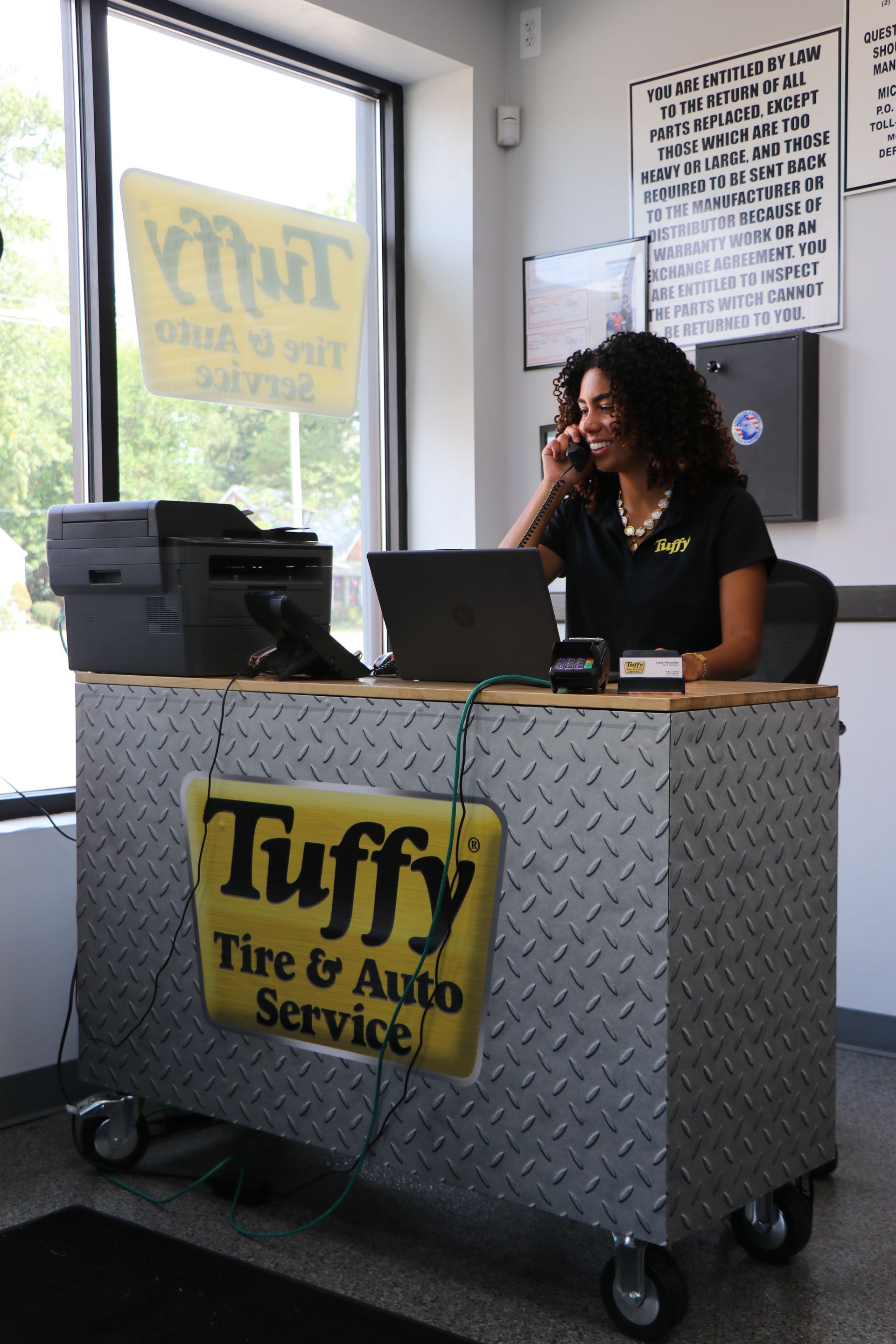 Woman on phone at a Tuffy Tire & Auto Service front desk, smiling, with a laptop, printer, and sign.