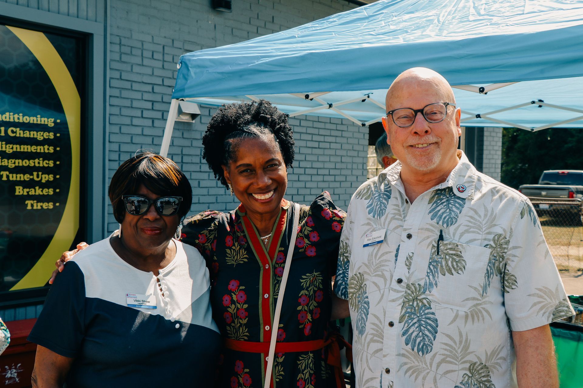 Three people smiling, posed outside near a light-blue tent and brick building.