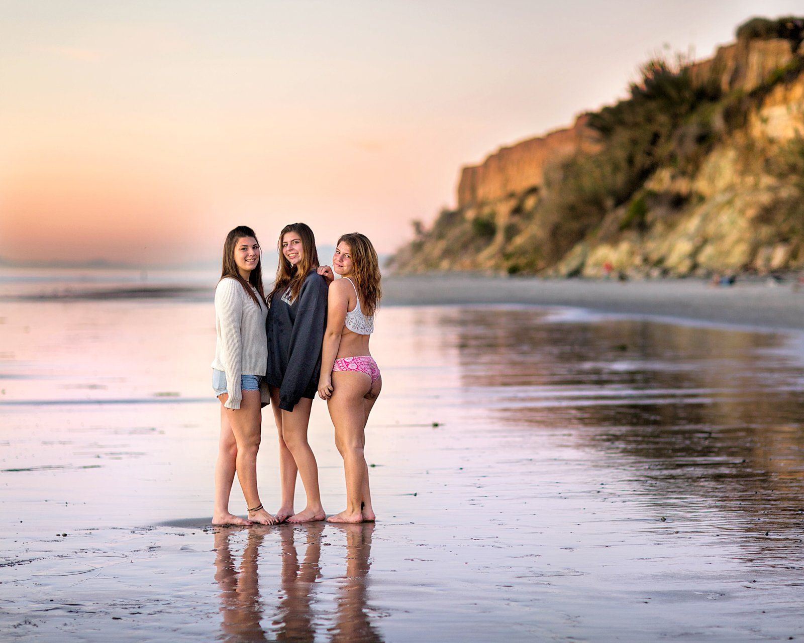Three young women are standing on a beach at sunset.