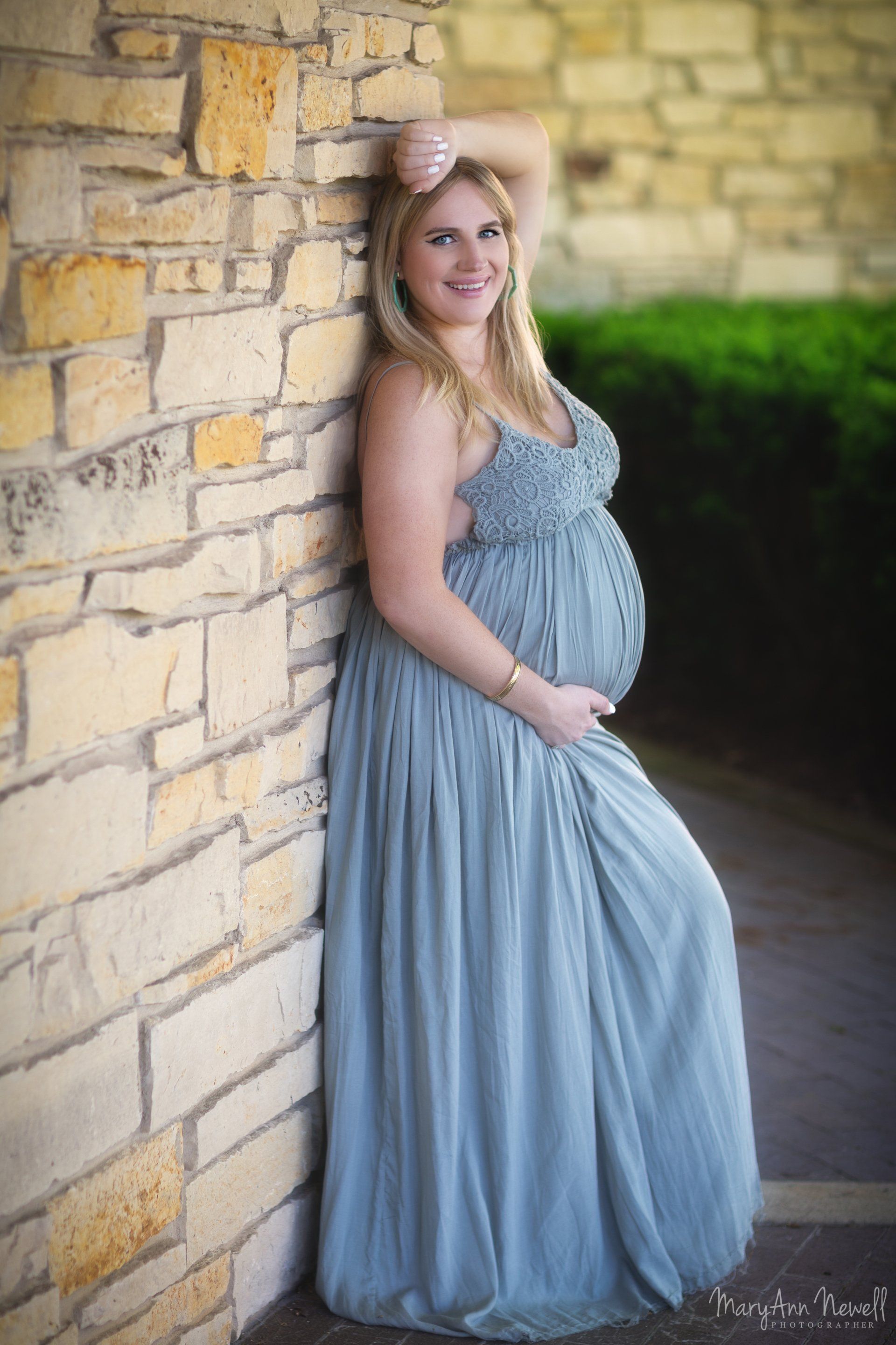 A pregnant woman in a blue dress is leaning against a stone wall.