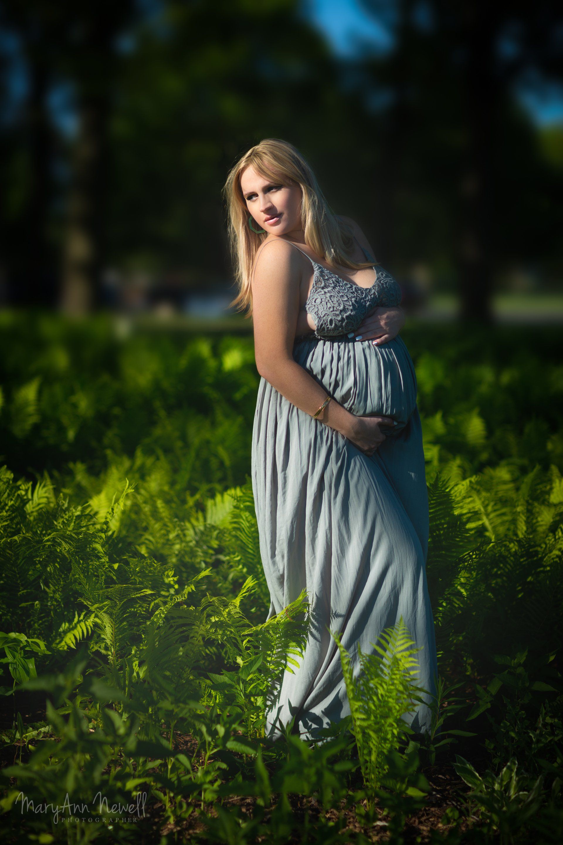 A pregnant woman is standing in a field of ferns holding her belly.