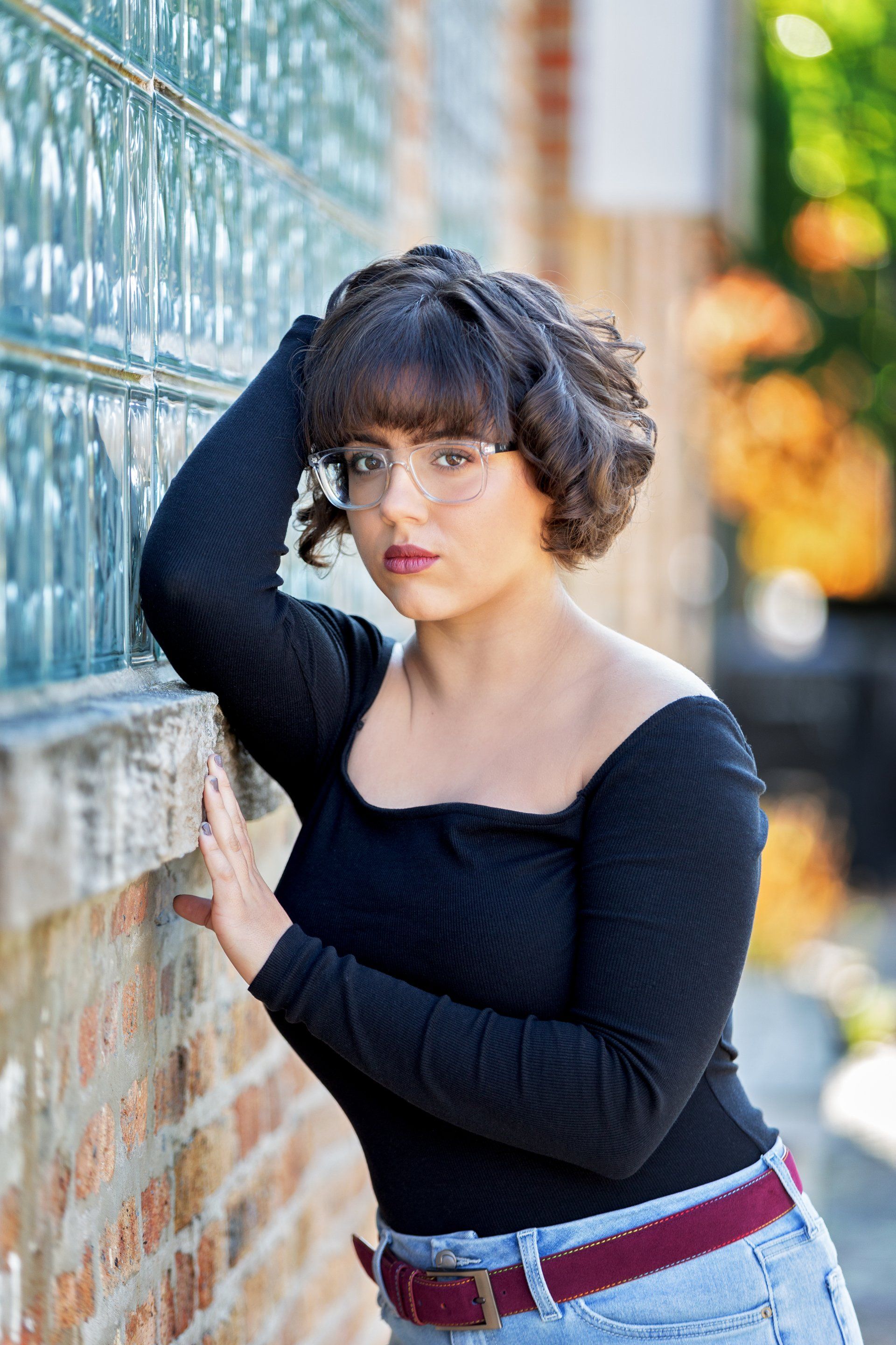 A woman wearing glasses and a black top is leaning against a brick wall.