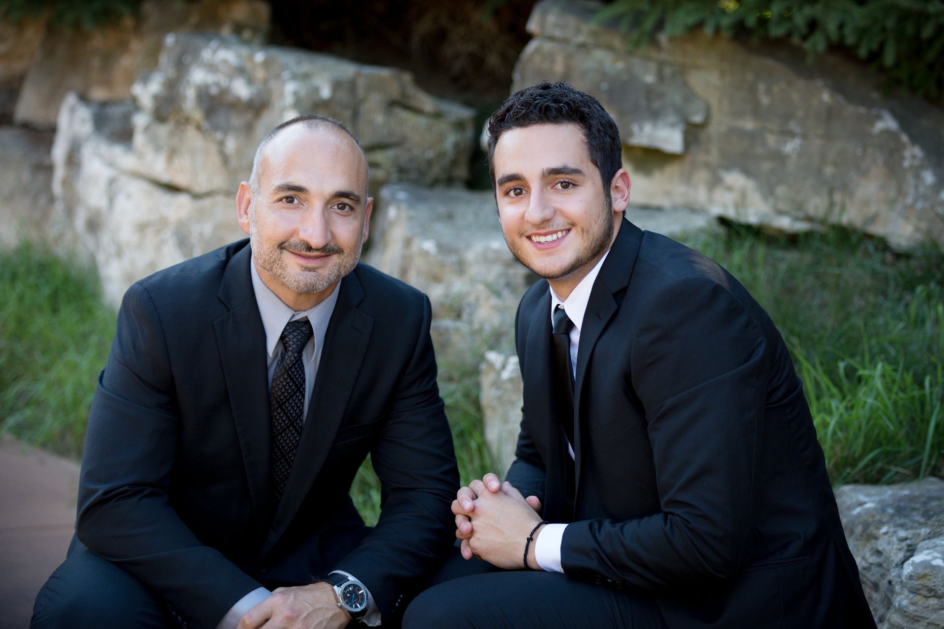 Two men in suits and ties are sitting next to each other on a rock.