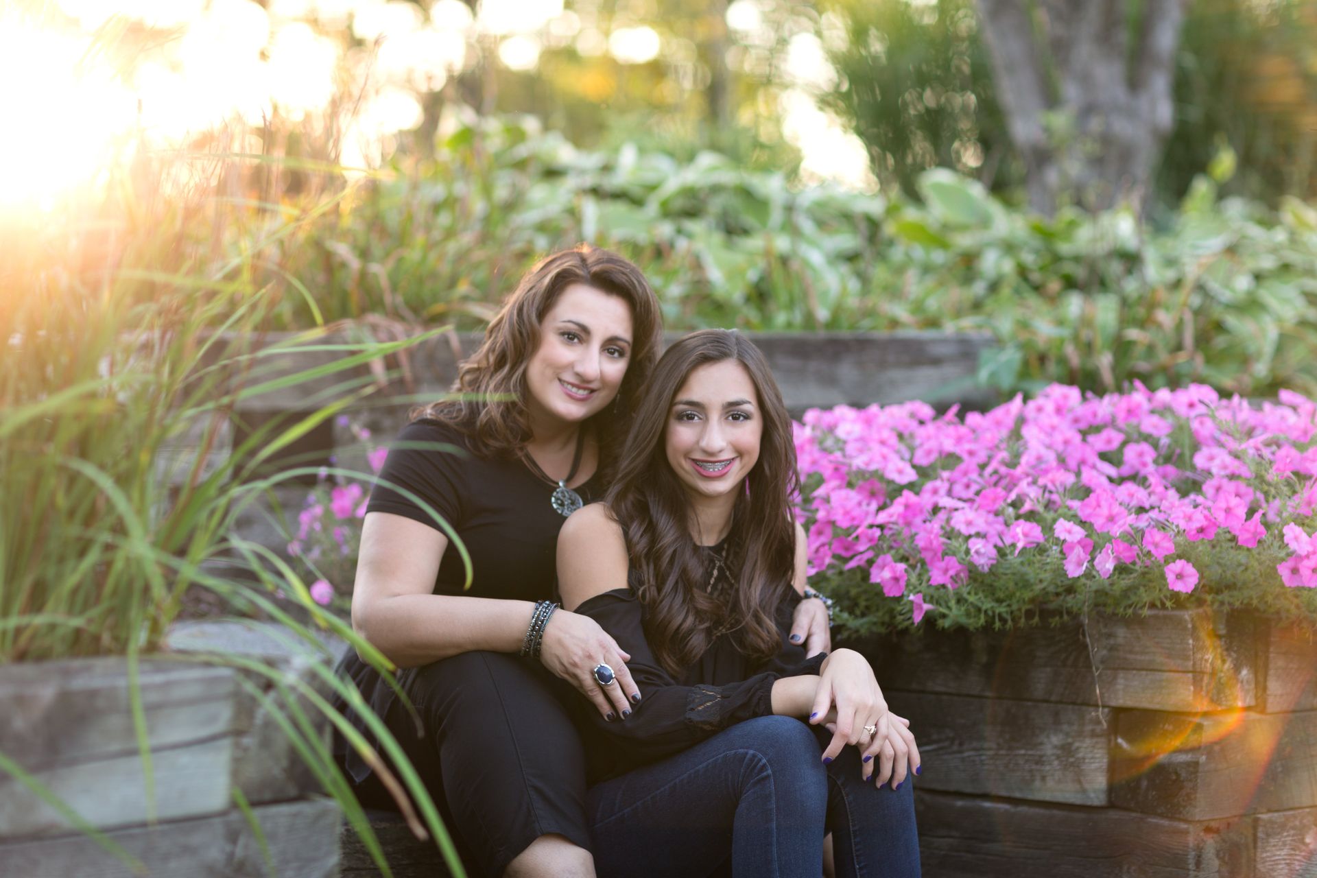 Two women are sitting next to each other on a bench in front of pink flowers.