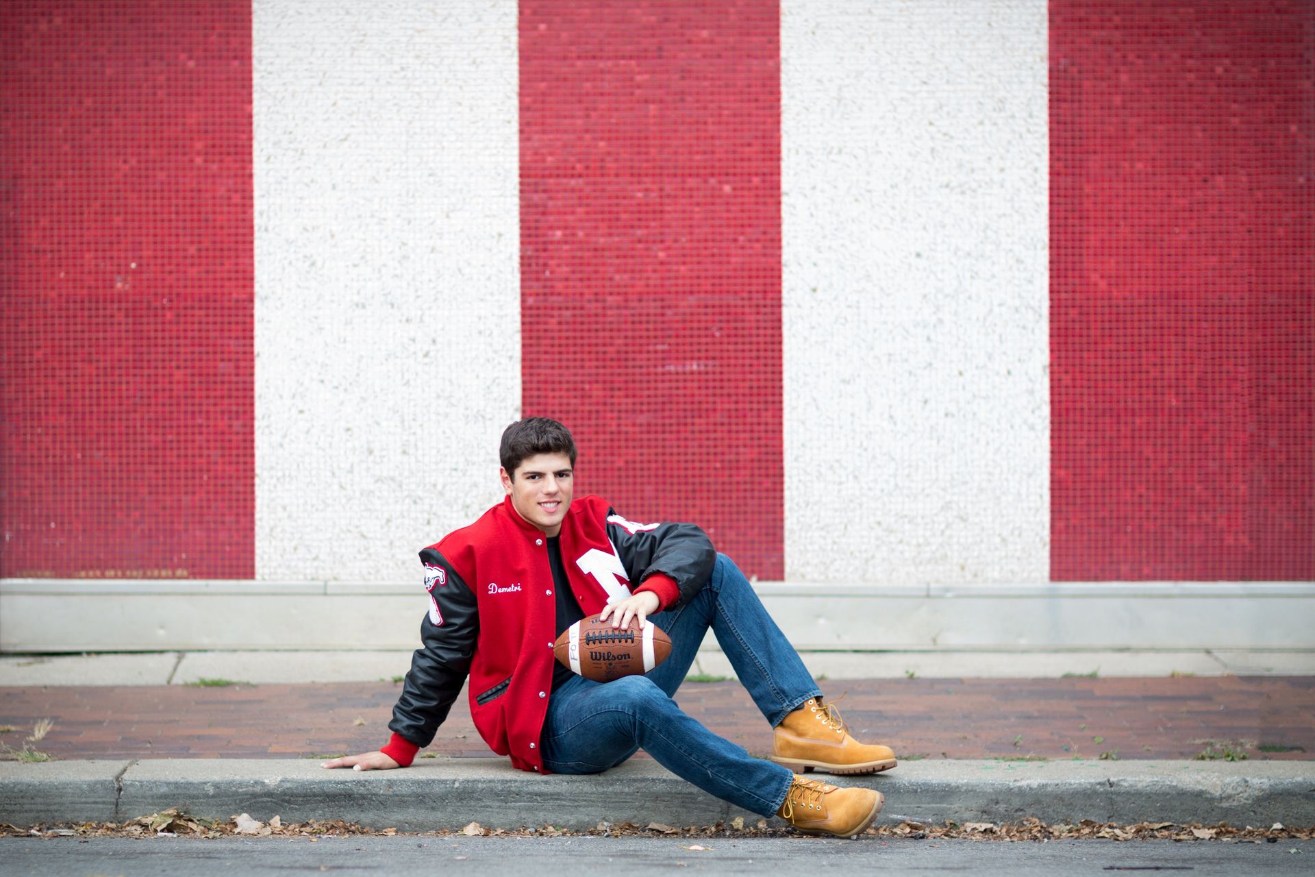 A young man is sitting on the sidewalk holding a football.