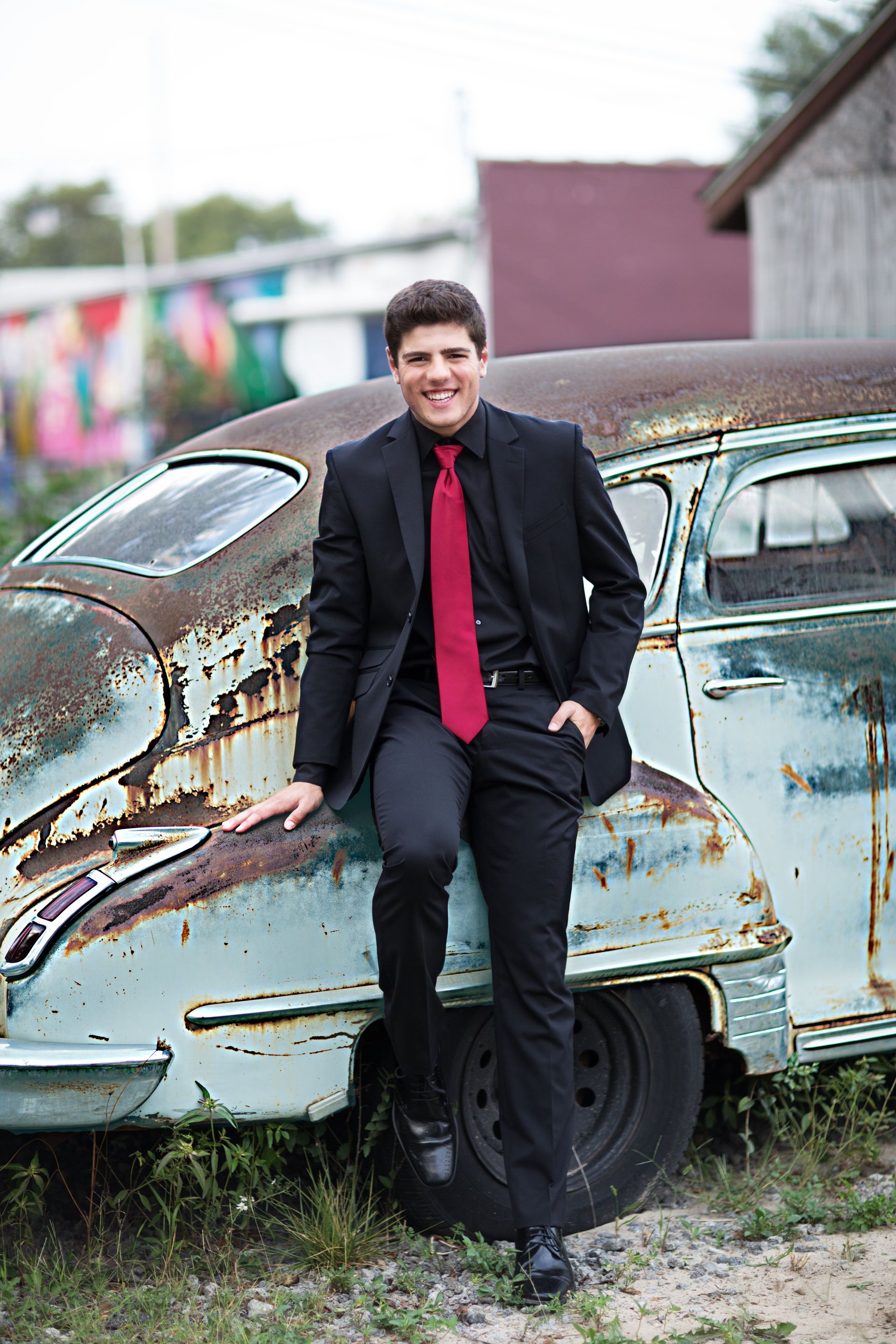 A man in a suit and tie is leaning against an old rusty car.