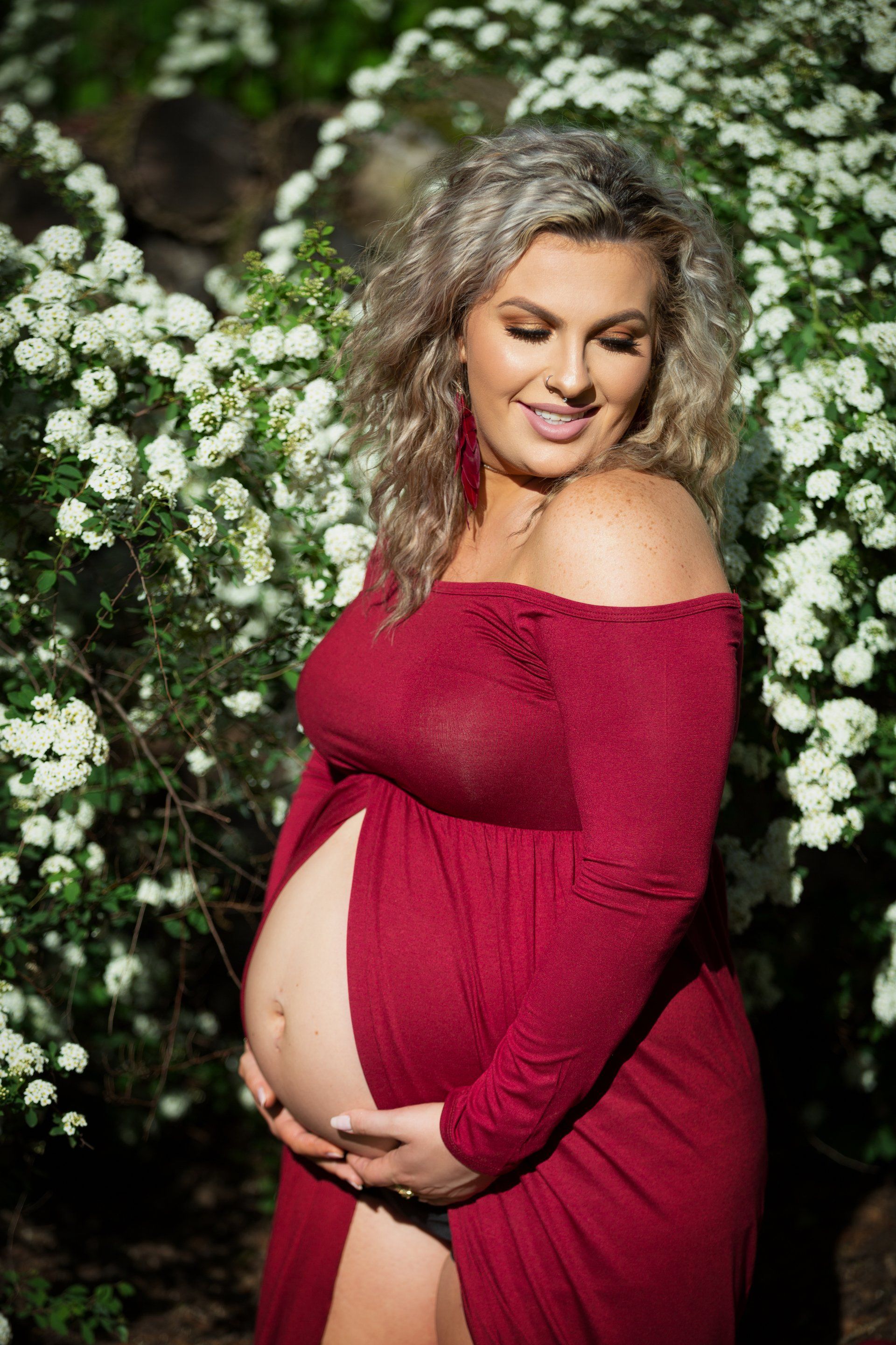 A pregnant woman in a red dress is standing in front of a bush with white flowers.