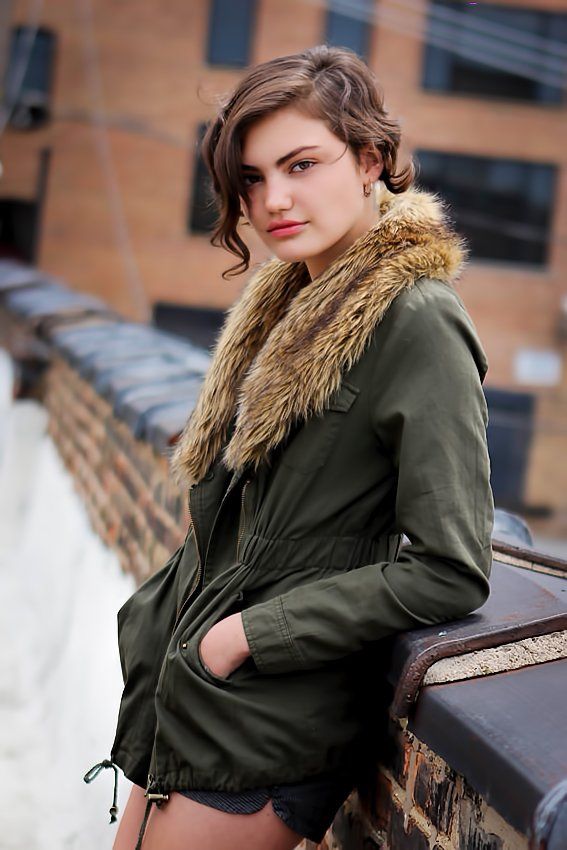 A woman in a green jacket with a fur collar is leaning against a wall.