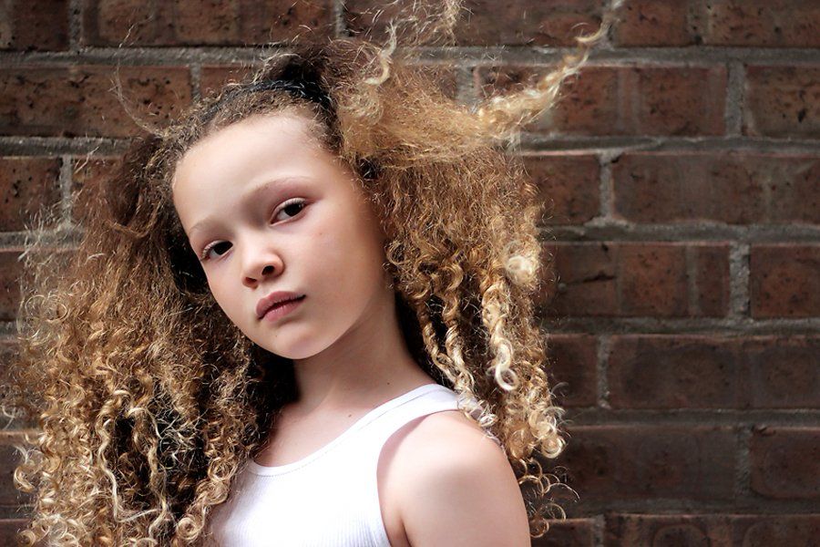 A young girl with curly hair is standing in front of a brick wall.