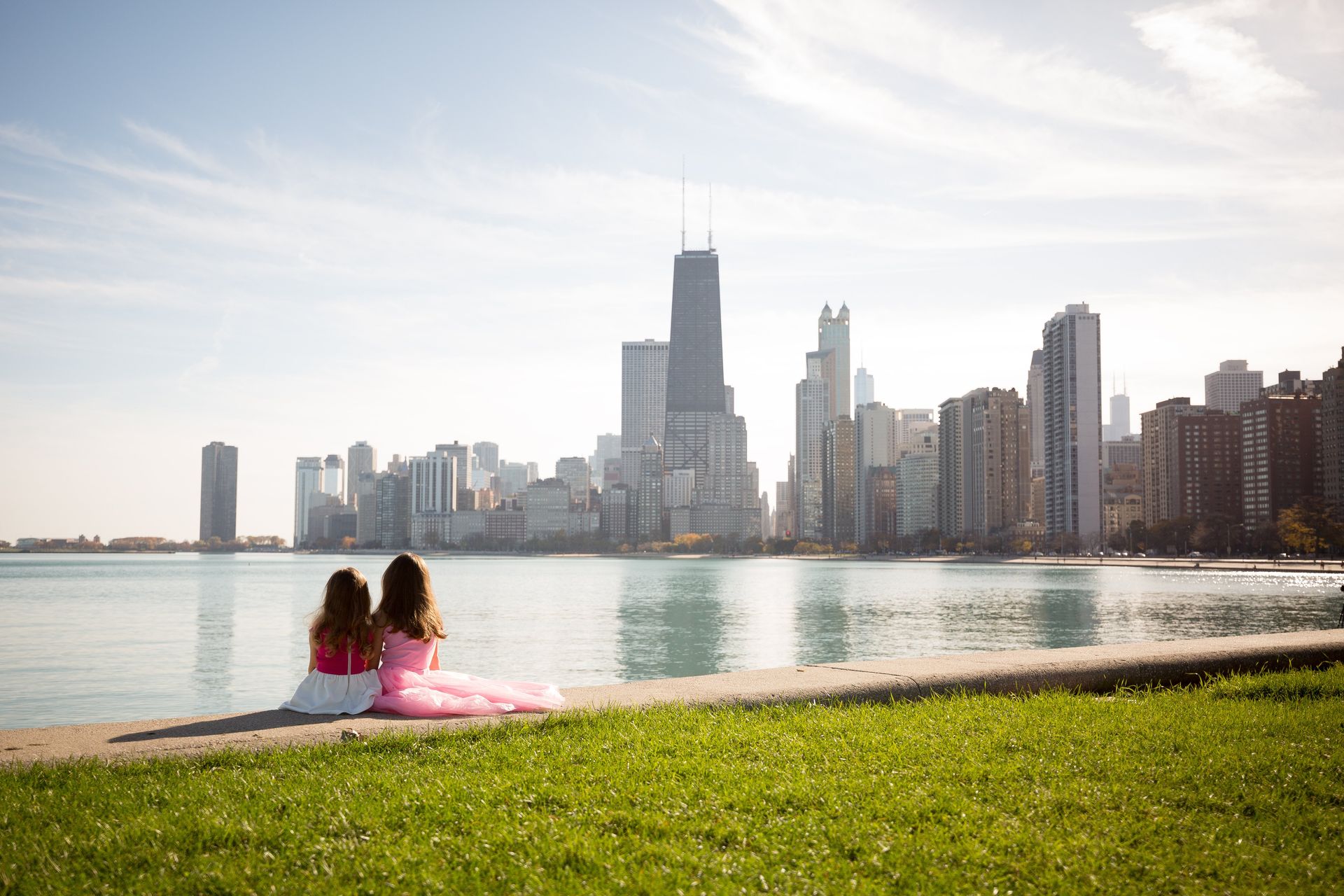 Two women are sitting on the shore of a lake looking at the city skyline.