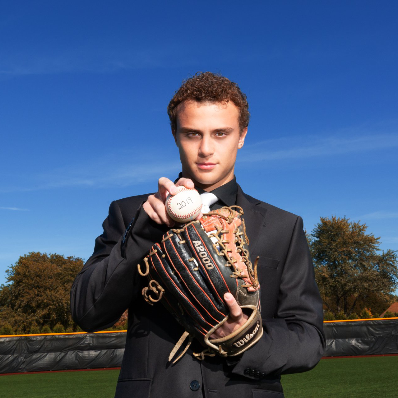 A man in a suit is holding a baseball and a glove with the letter s on it