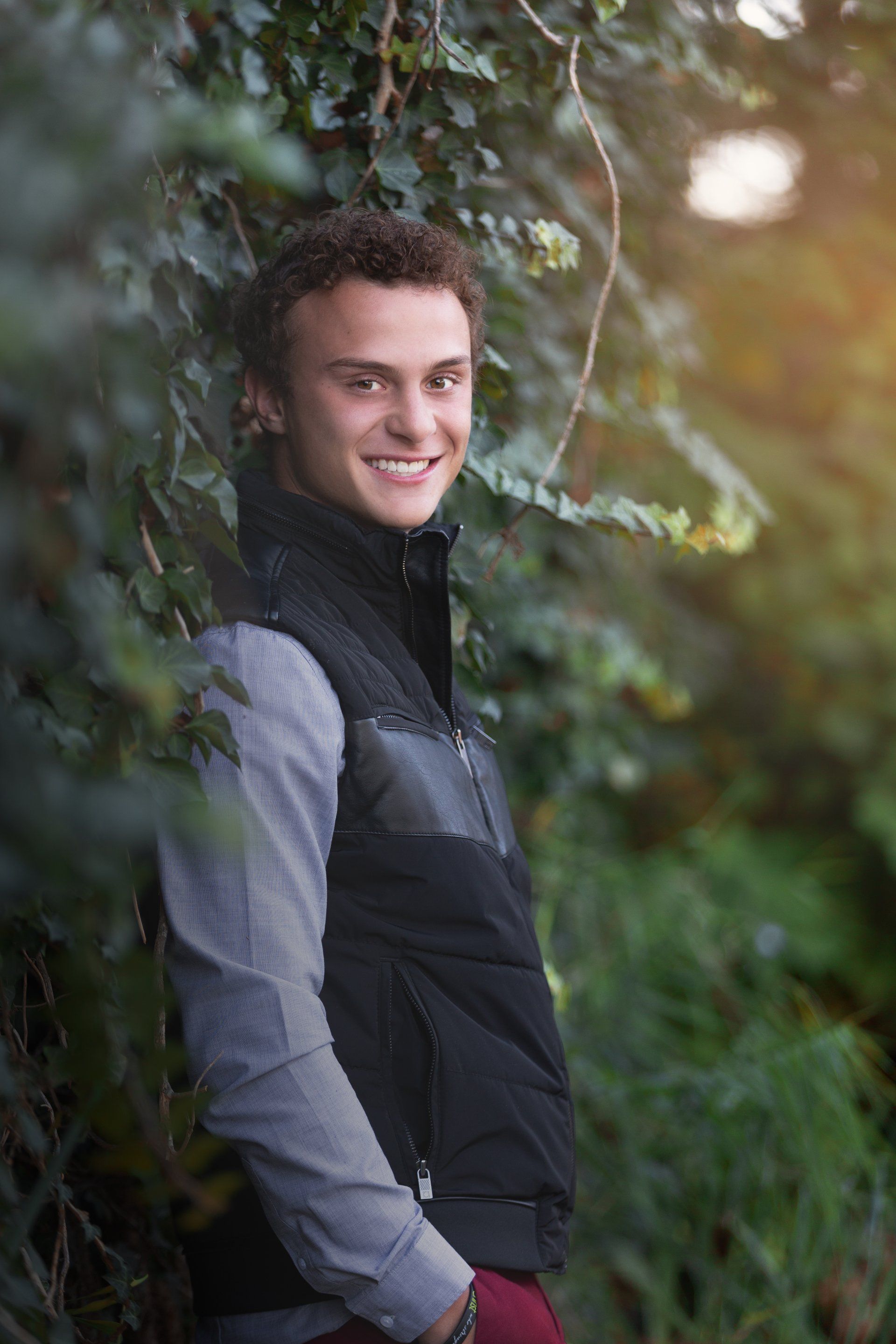 A young man is leaning against a tree in the woods and smiling.