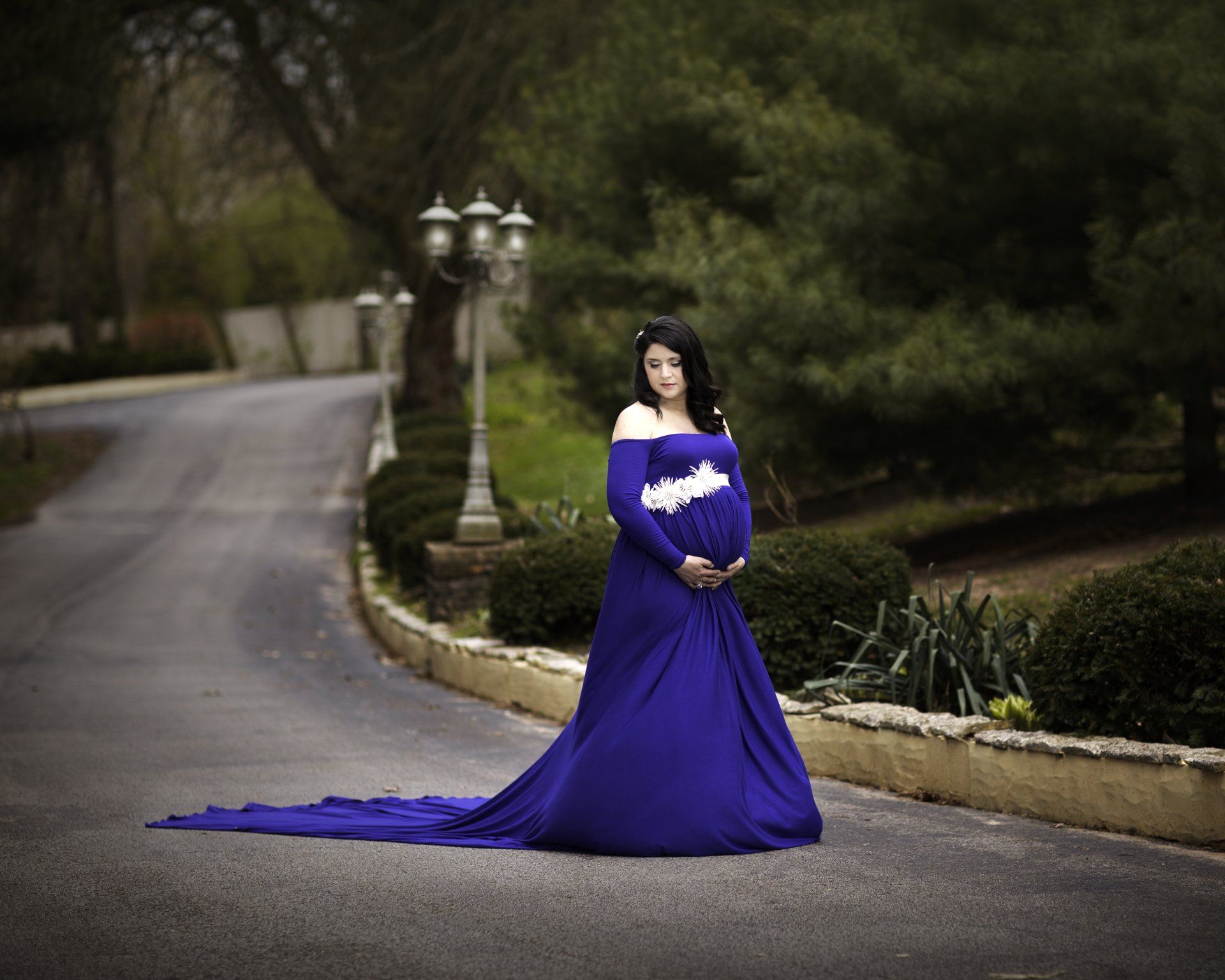 A pregnant woman in a long blue dress is standing on the side of a road.