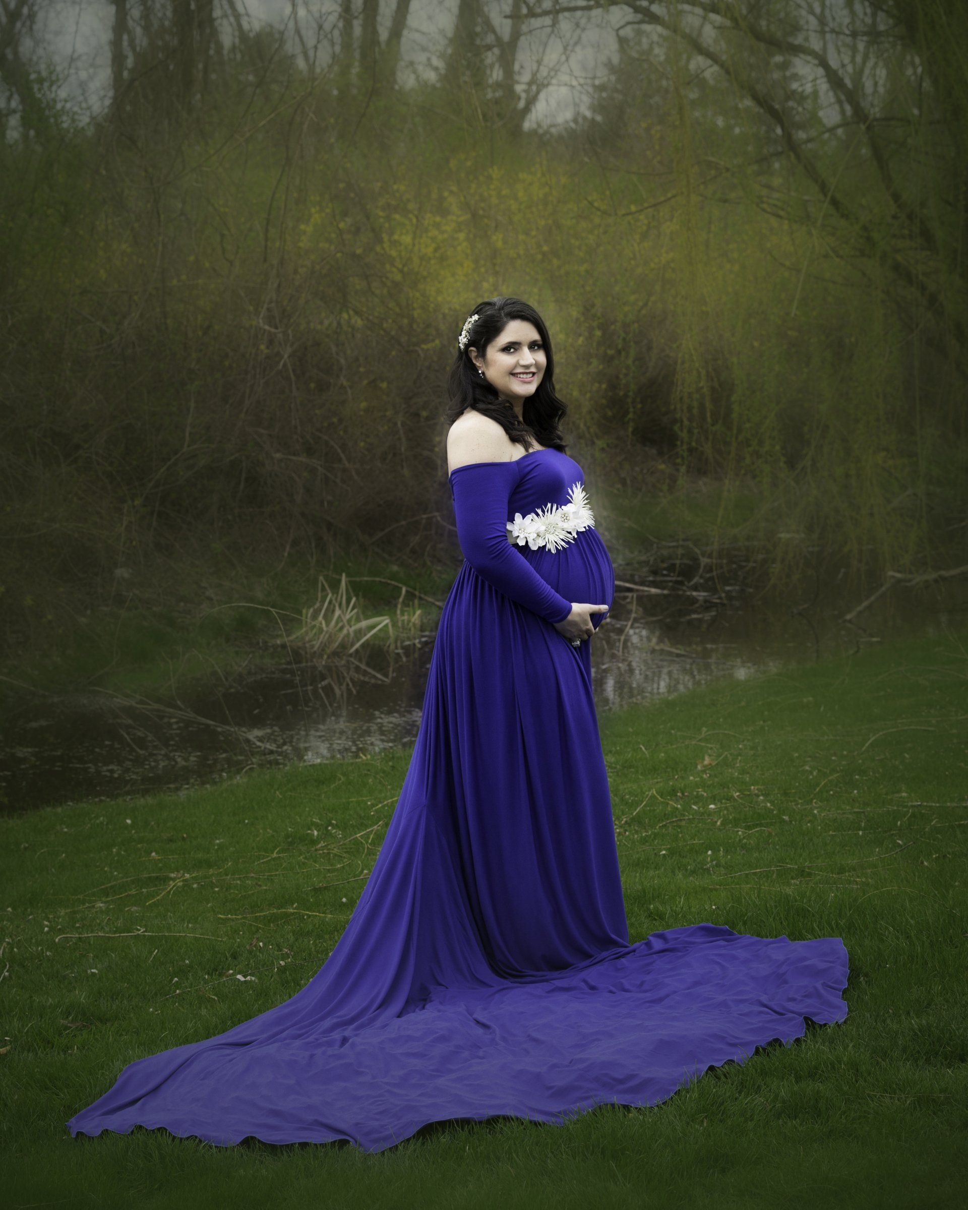 A pregnant woman in a long purple dress is standing in a field.