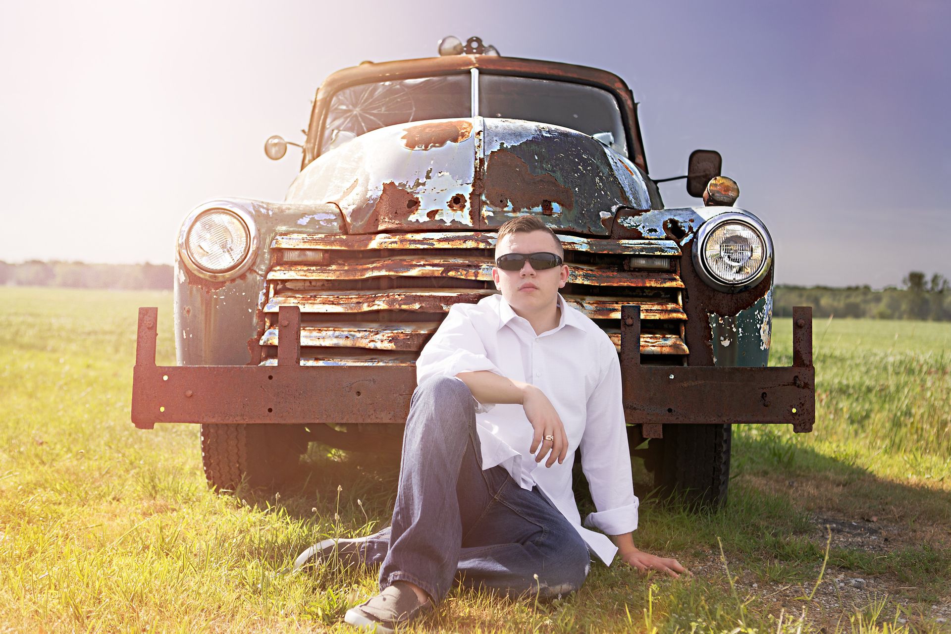 A man is kneeling in front of a rusty truck in a field.
