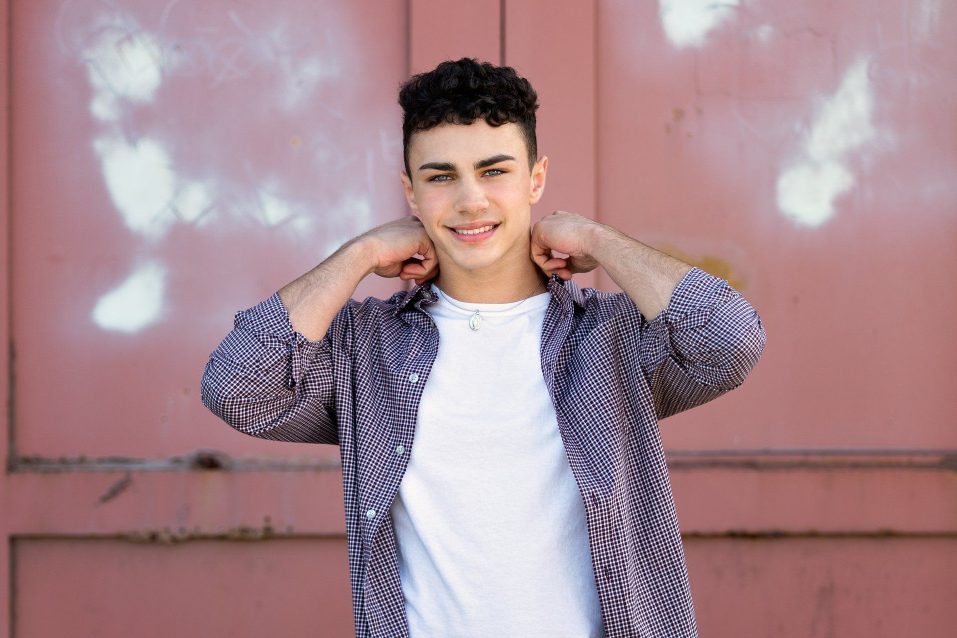 A young man wearing a plaid shirt and a white t-shirt is standing in front of a red wall.