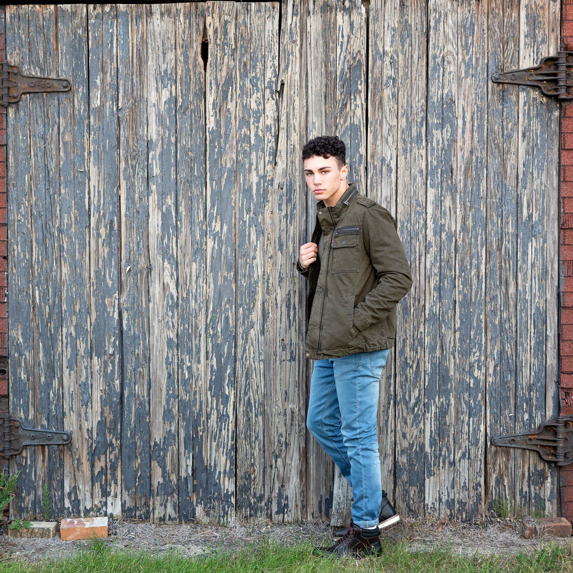 A young man in a green jacket is standing in front of a wooden door.