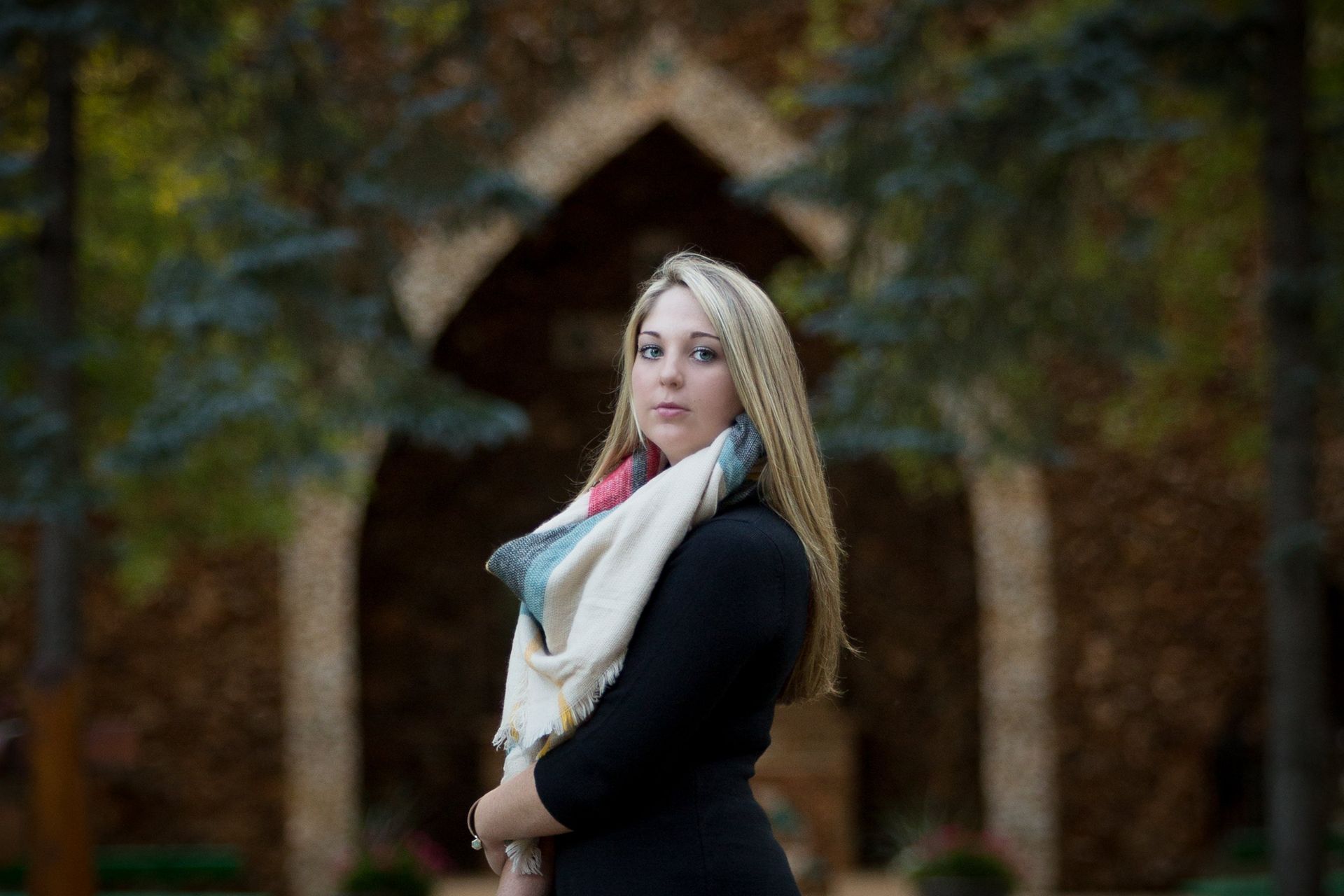A woman wearing a scarf and a black dress is standing in front of a stone archway.