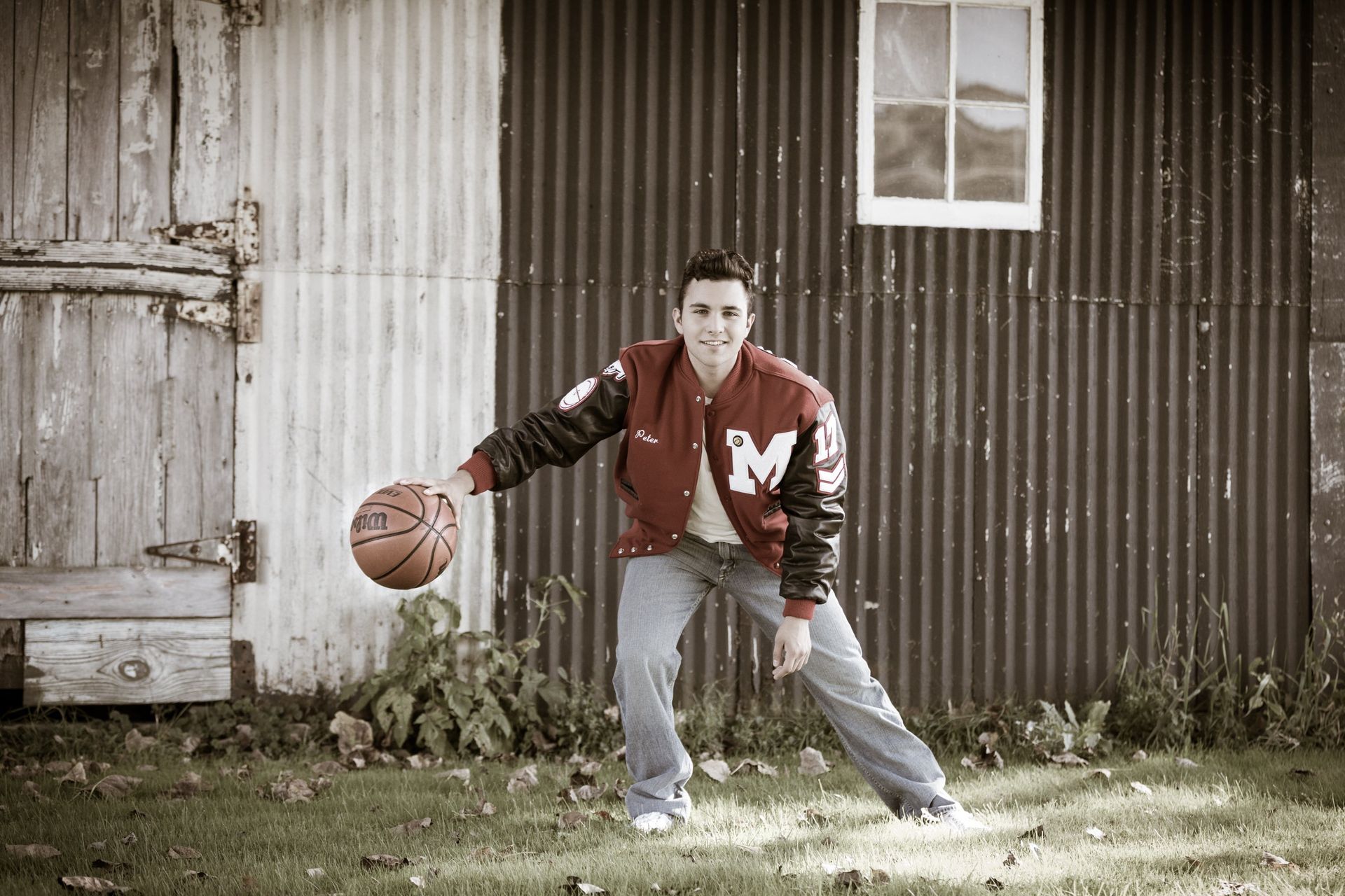 A man in a letterman jacket is holding a basketball in front of a barn.