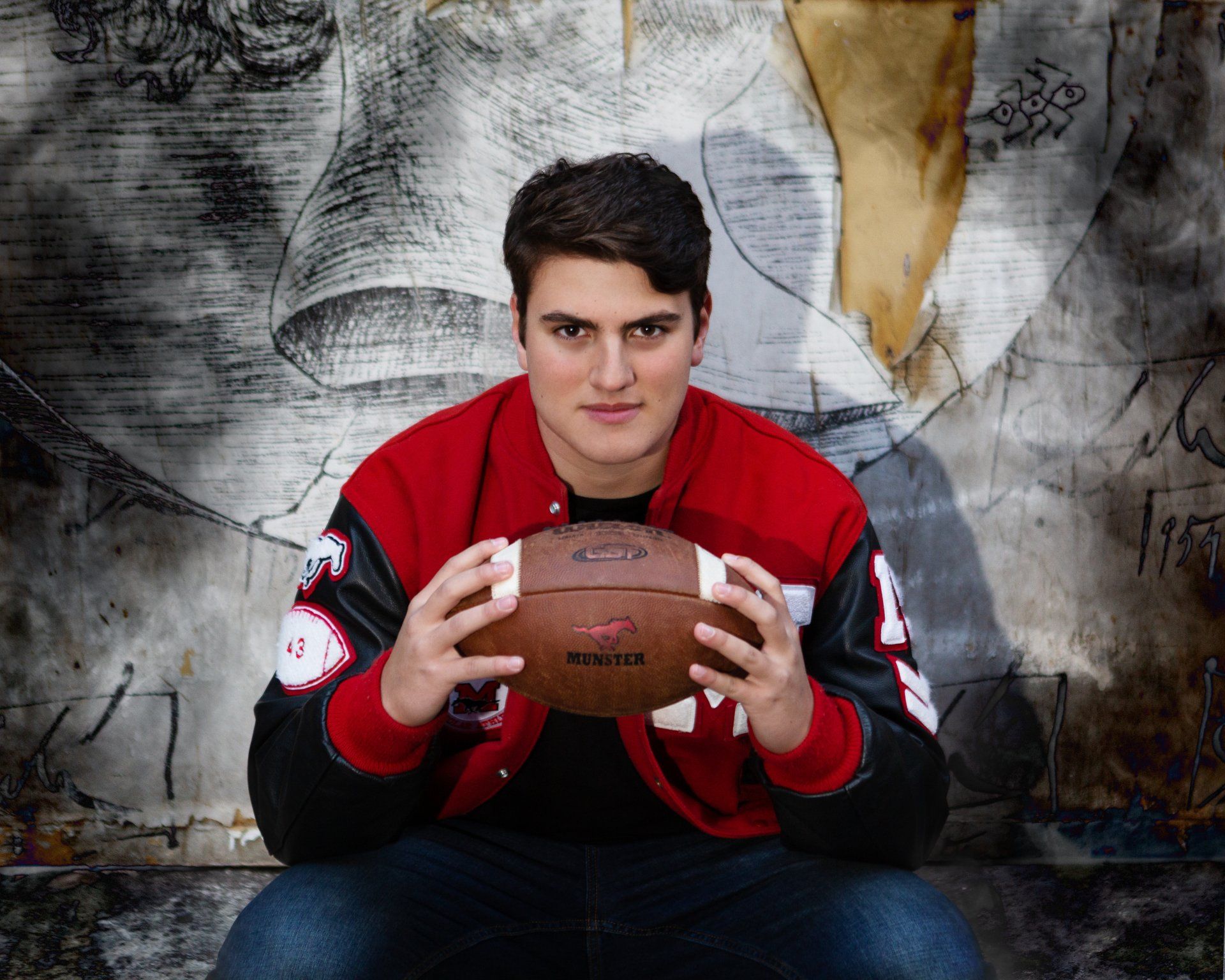 A young man in a red jacket is holding a football in front of a graffiti wall.