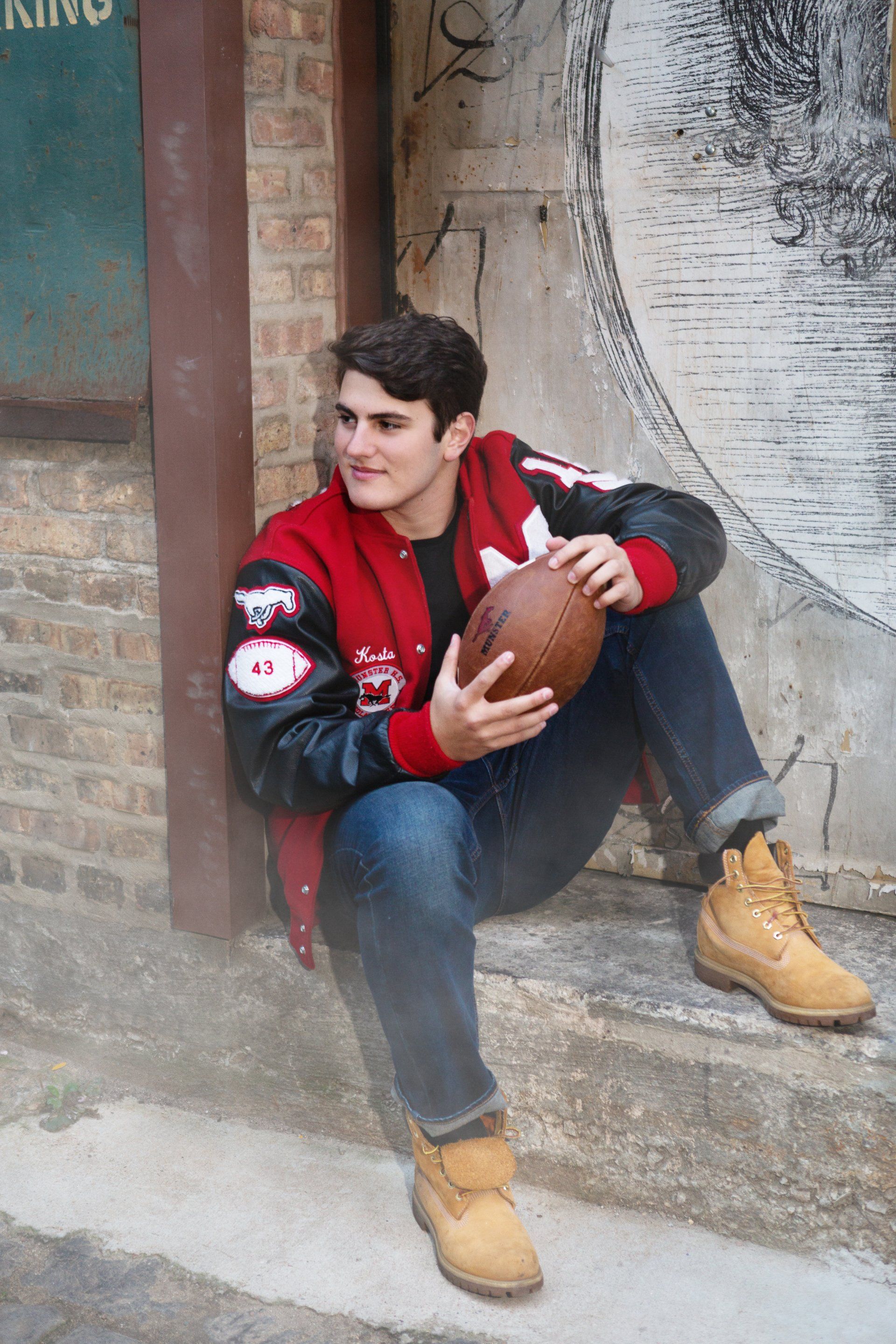 A man in a varsity jacket is sitting on the steps holding a football.