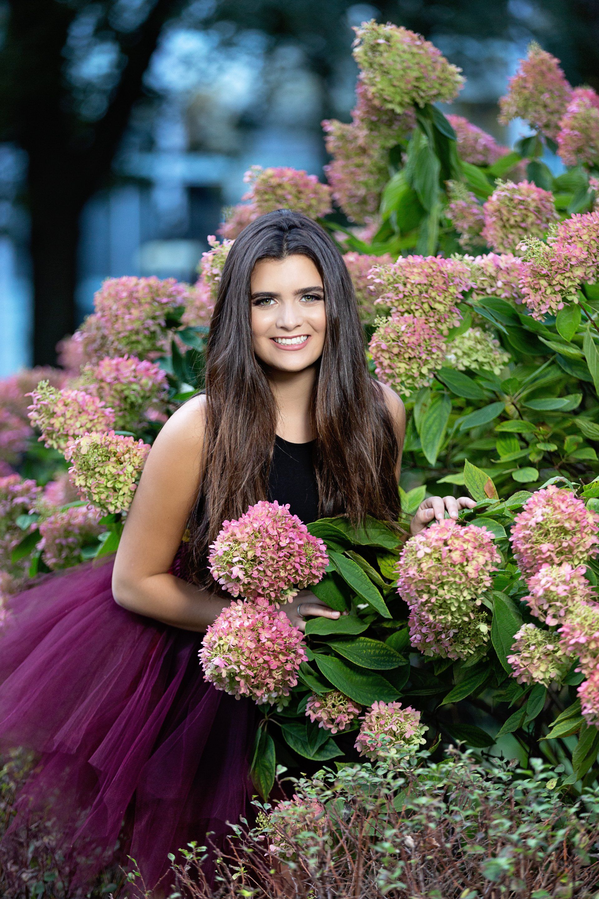 A woman in a purple skirt is standing next to a bush of pink flowers.
