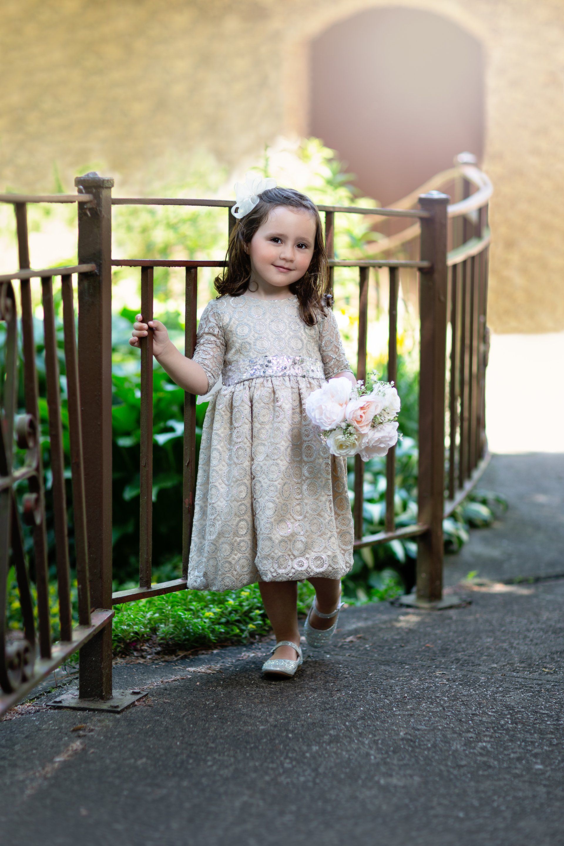 A little girl in a dress is standing next to a fence holding a bouquet of flowers.