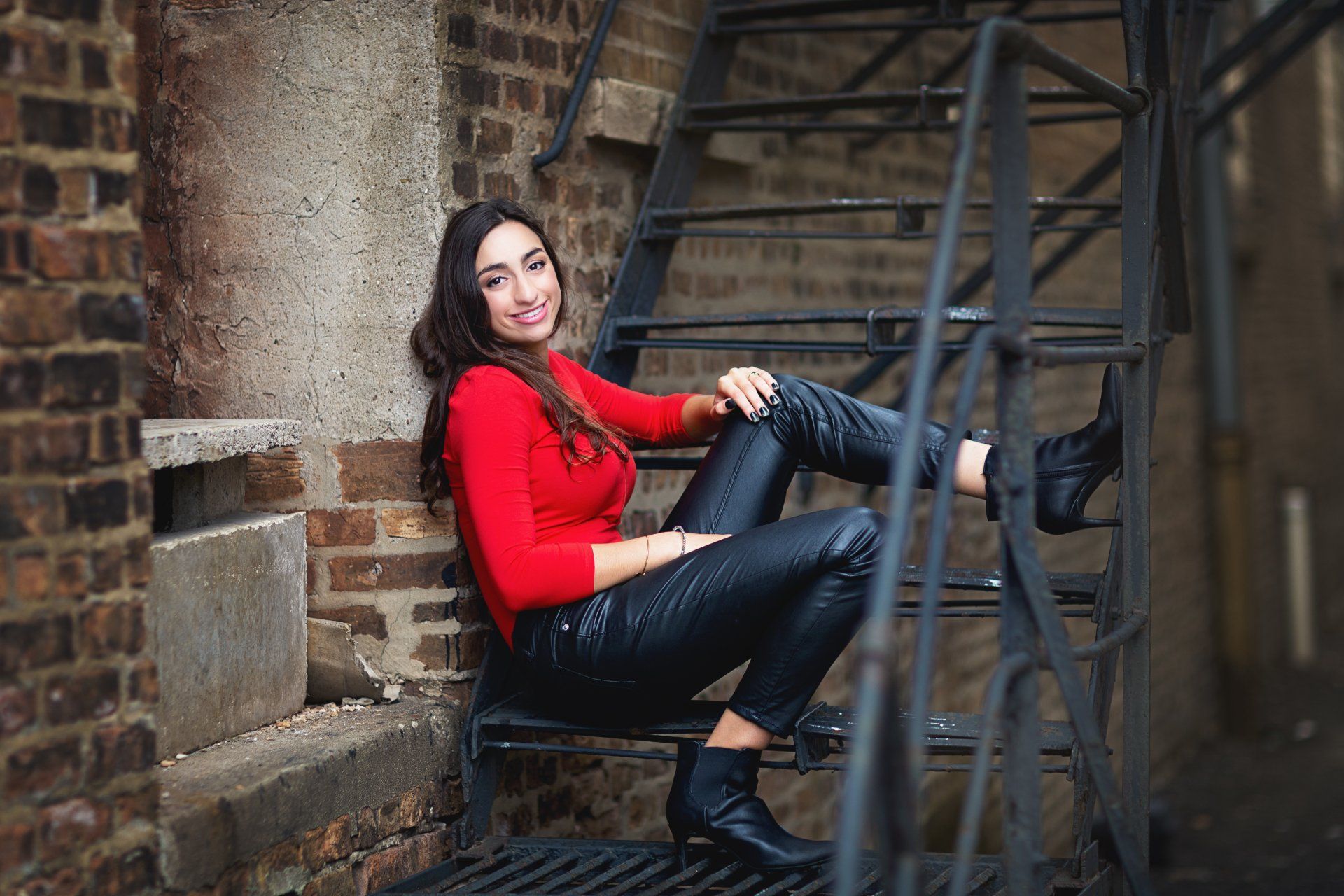 A woman in a red sweater and black pants is sitting on a fire escape.