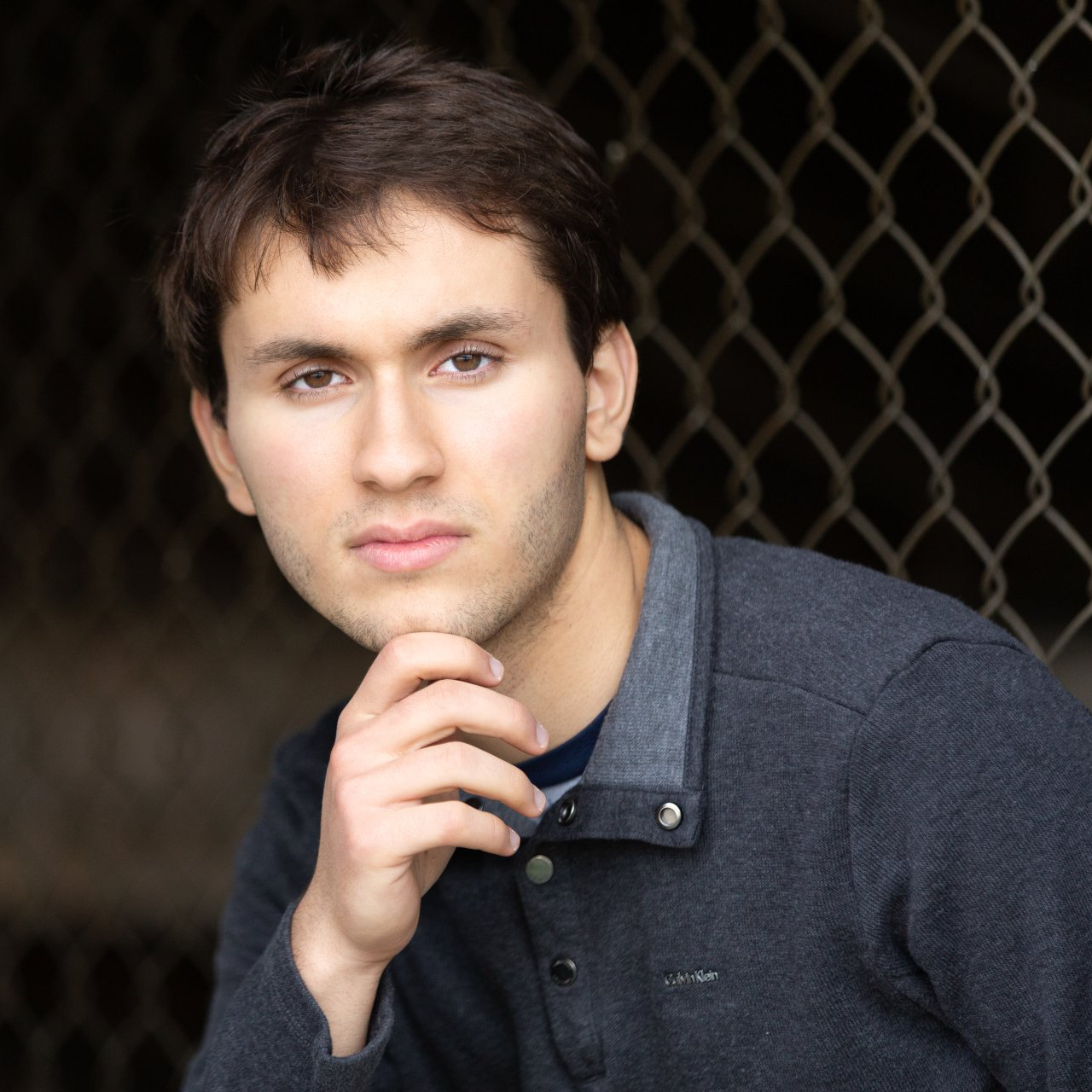 A man in a grey sweater is sitting in front of a chain link fence