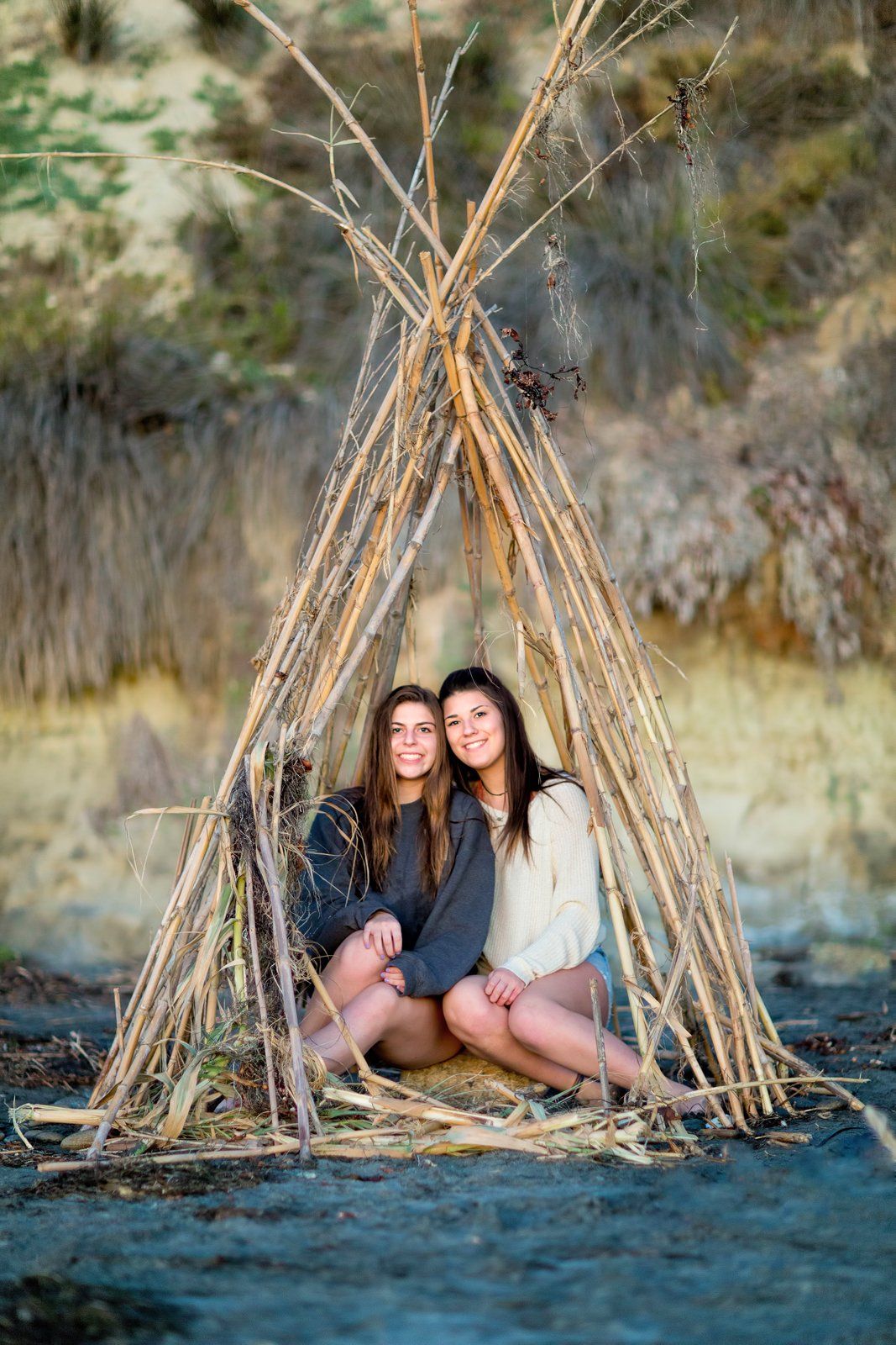 Two girls are sitting in a teepee made of sticks on the beach.