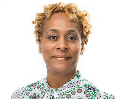 A woman with curly hair is wearing a floral shirt and smiling.