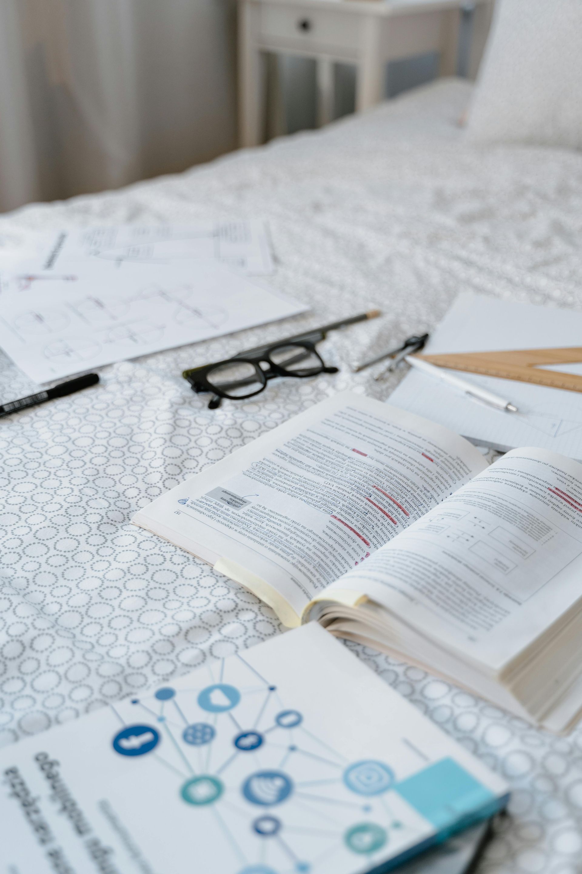 Book laid open on a bed with glasses, paper, pens and pencills