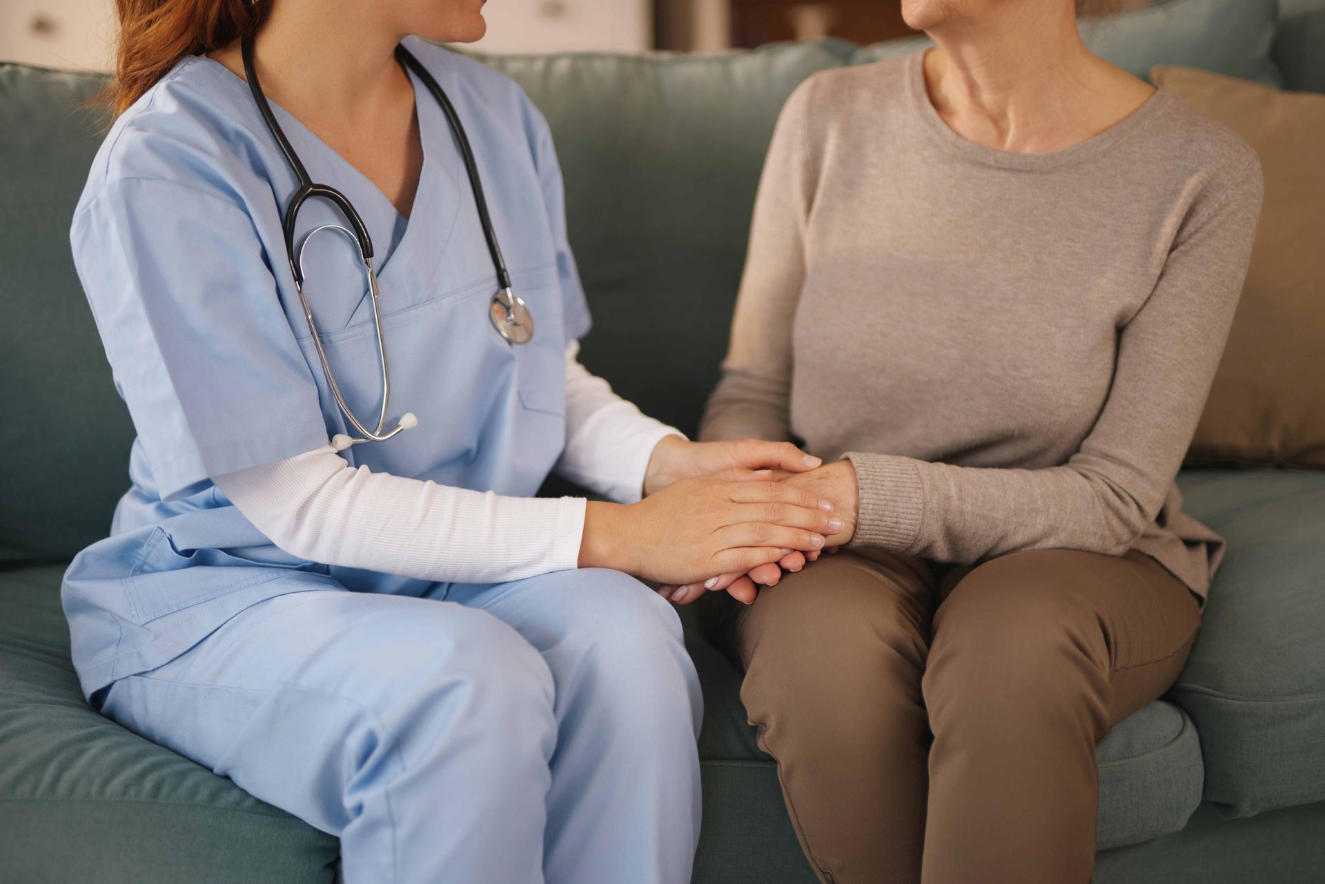 Nurse in blue scrubs holding hands with a patient on a couch.